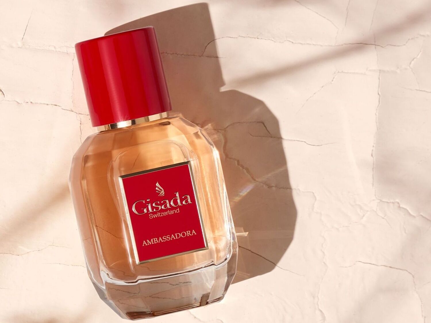 Šveicariškas „Gisada Ambassadora“ aromatas skaidriame stikliniame buteliuke su raudonu dangteliu ir etikete ant smėlio spalvos paviršiaus.