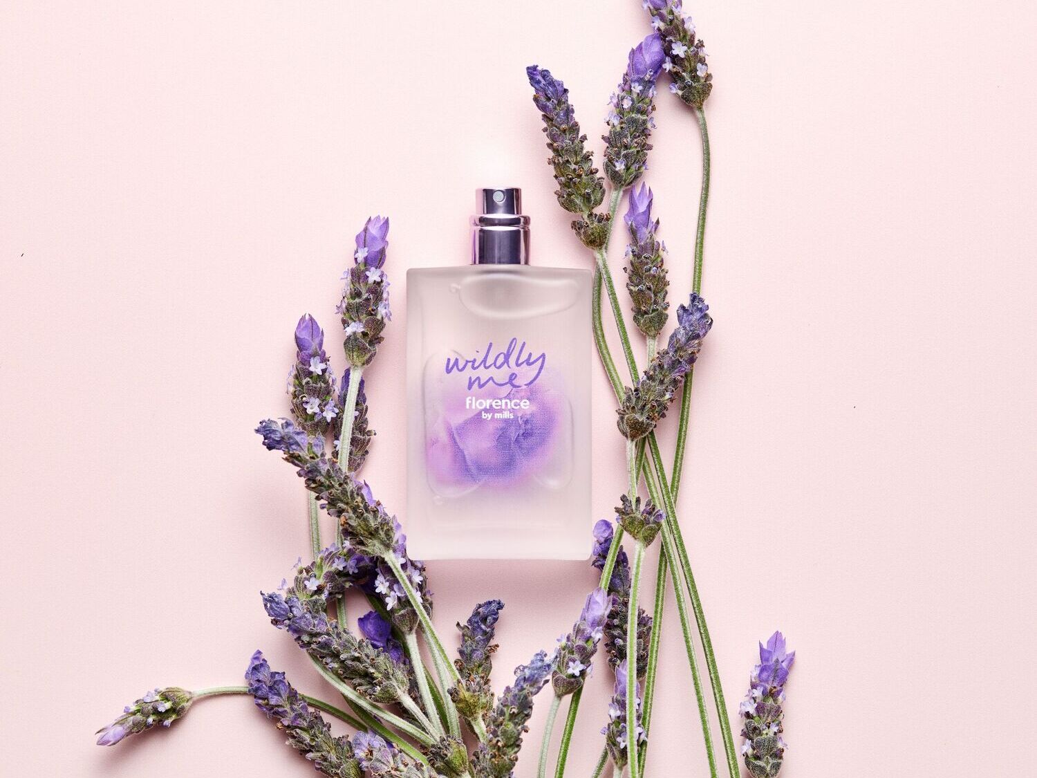 Švelniai rožiniame fone pavaizduotas „Florence by Mills Wildly Me“ aromatas, apsuptas levandų šakelių, kurios pabrėžia jo gaivumą ir natūralumą.
