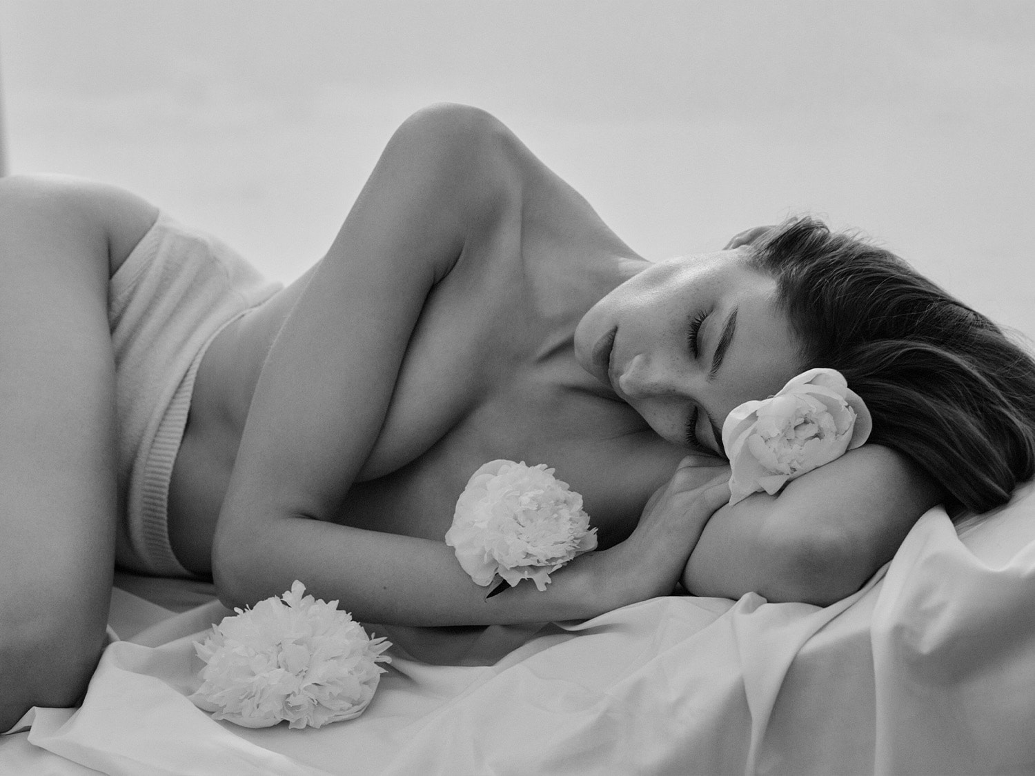 En noir et blanc, une femme se repose, les yeux clos, entourée de pivoines blanches, évoquant la douceur et la sérénité.