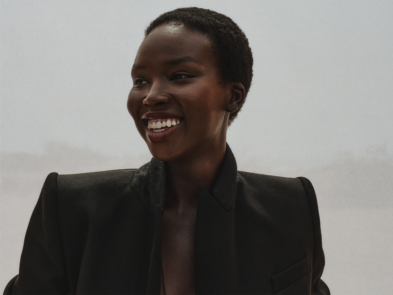 Portrait d'une femme souriante à la peau foncée, portant un blazer noir élégant, regardant vers la gauche avec une expression joyeuse.