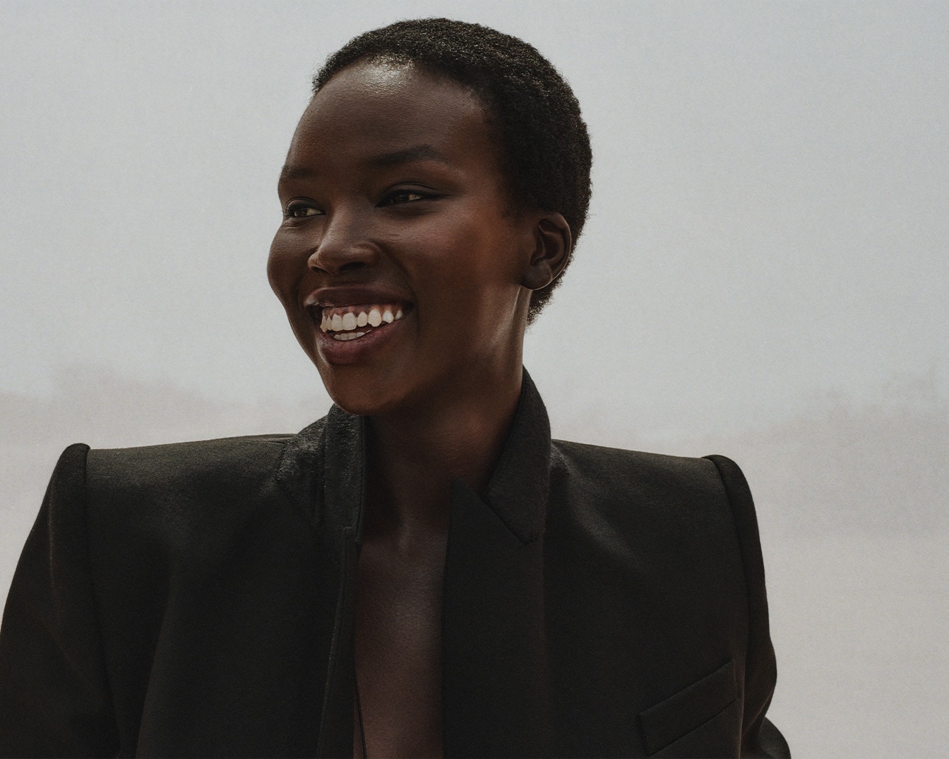 Švytinti juodaodė moteris su trumpais plaukais ir plačia šypsena, vilkinti juodą švarką, spinduliuoja pasitikėjimą ir eleganciją.