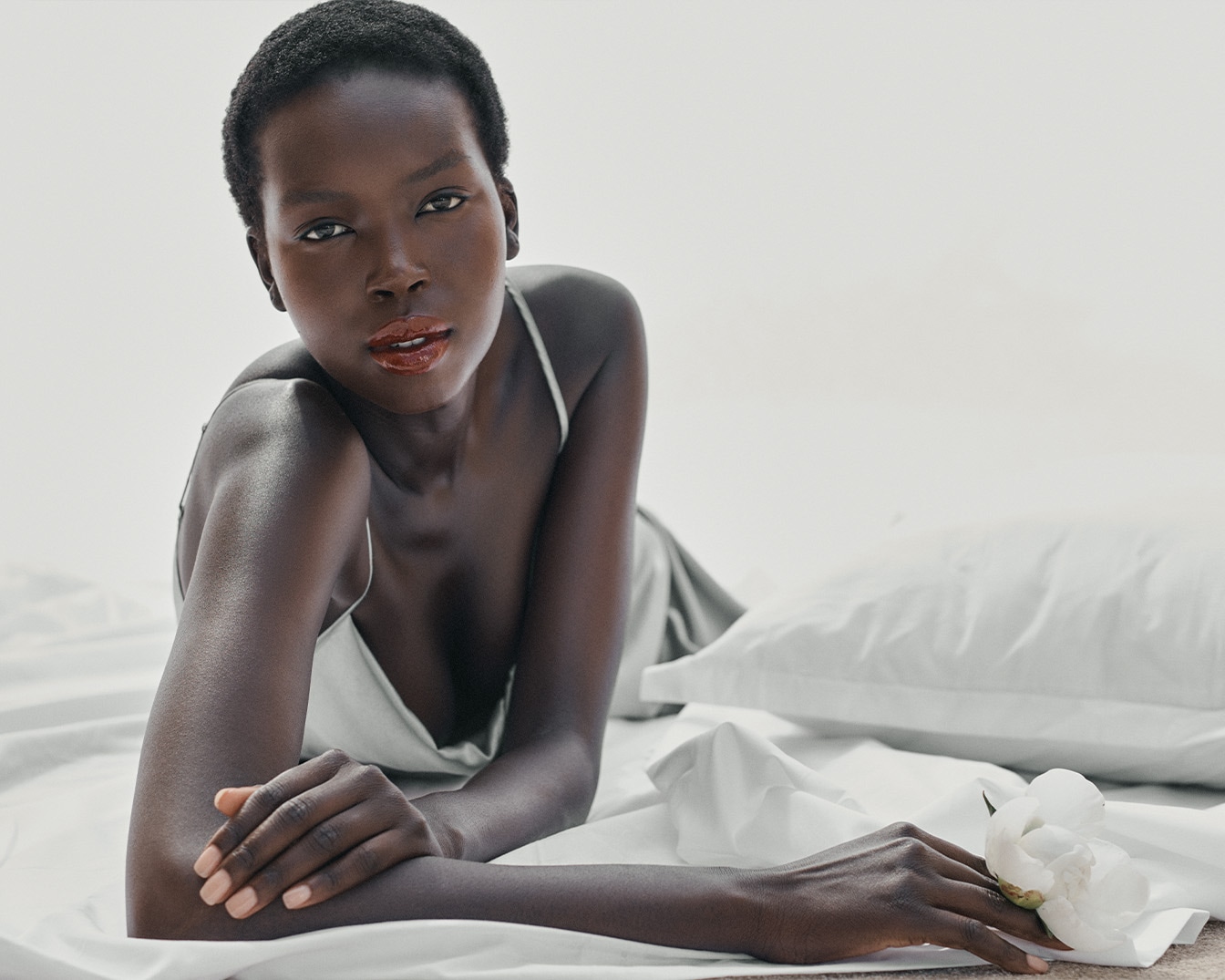 Tamsiaodė moteris su šviesia suknele guli ant baltos patalynės, šalia baltas žiedas, jos lūpos blizga.