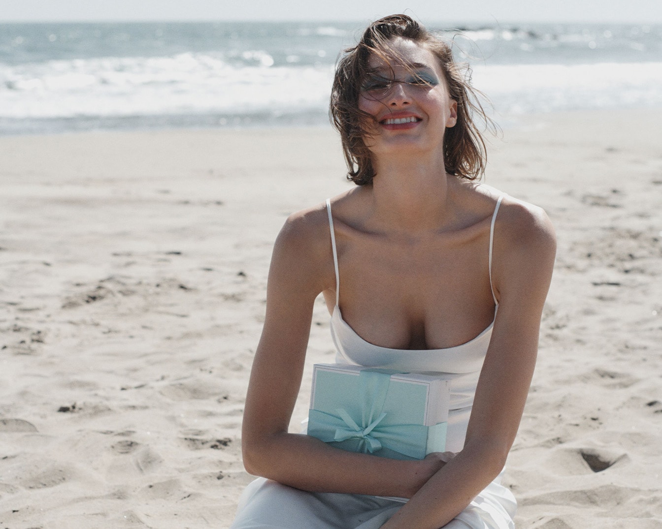 Sur la plage, une femme souriante en robe blanche tient un cadeau bleu clair, évoquant la joie des moments précieux.