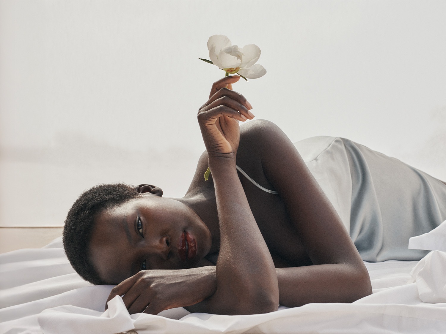 Une femme est allongée sur un tissu blanc, tenant une fleur blanche, créant une ambiance de sérénité et de beauté naturelle.