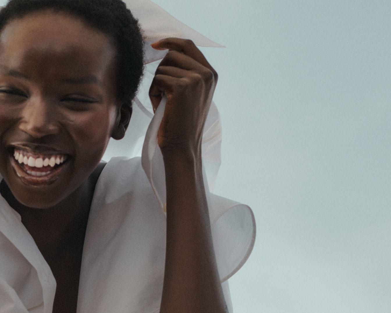 Une femme noire rayonnante, vêtue d'une chemise blanche, sourit en tenant un tissu léger, évoquant la joie et la fraîcheur.