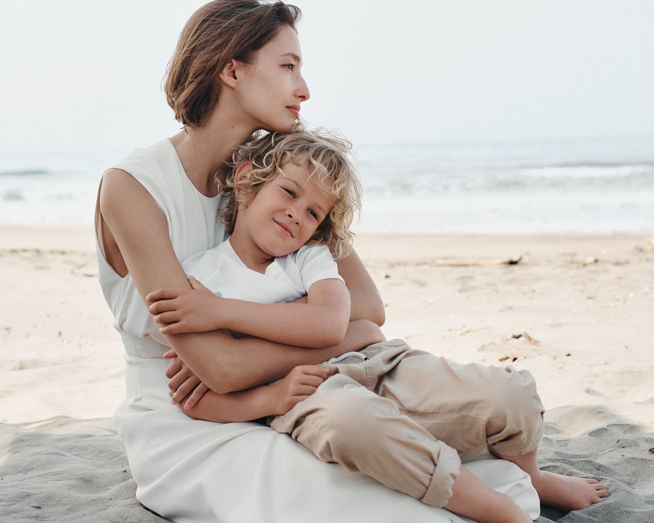 Sur la plage, une mère en robe blanche enlace tendrement son jeune fils aux cheveux bouclés, créant un moment de douceur et de connexion.