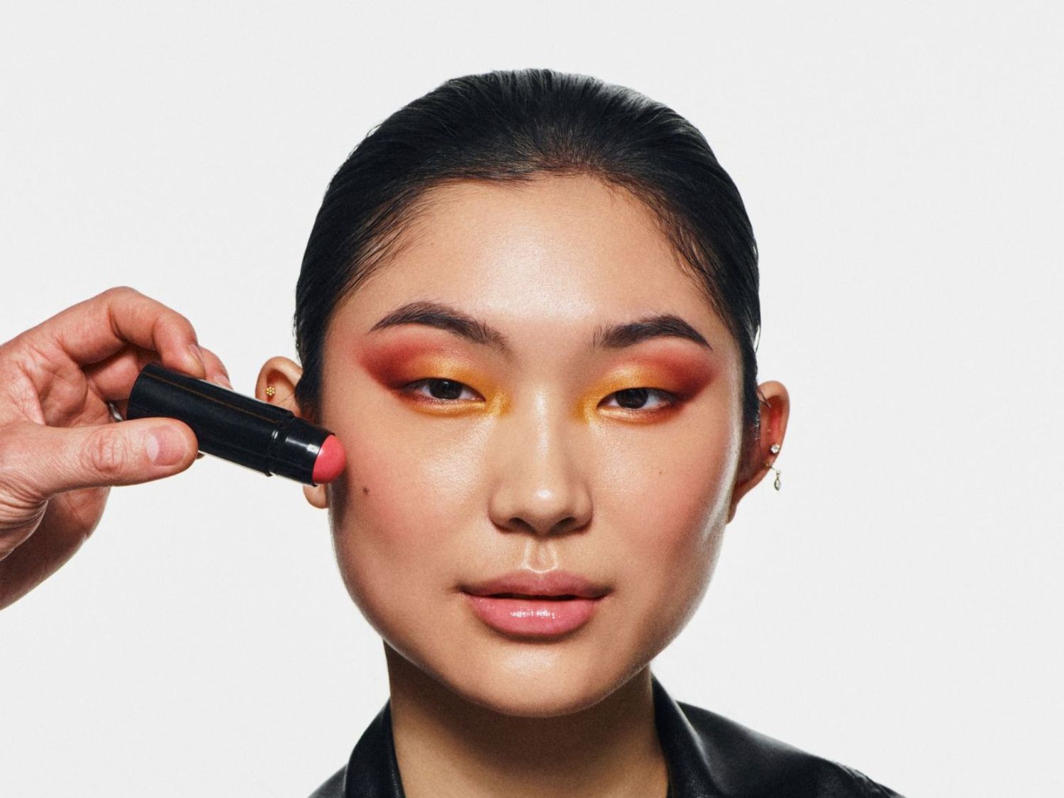 Un bâton de blush rose est appliqué sur la joue d'une femme au teint clair, avec un maquillage des yeux aux tons orange et rouge.