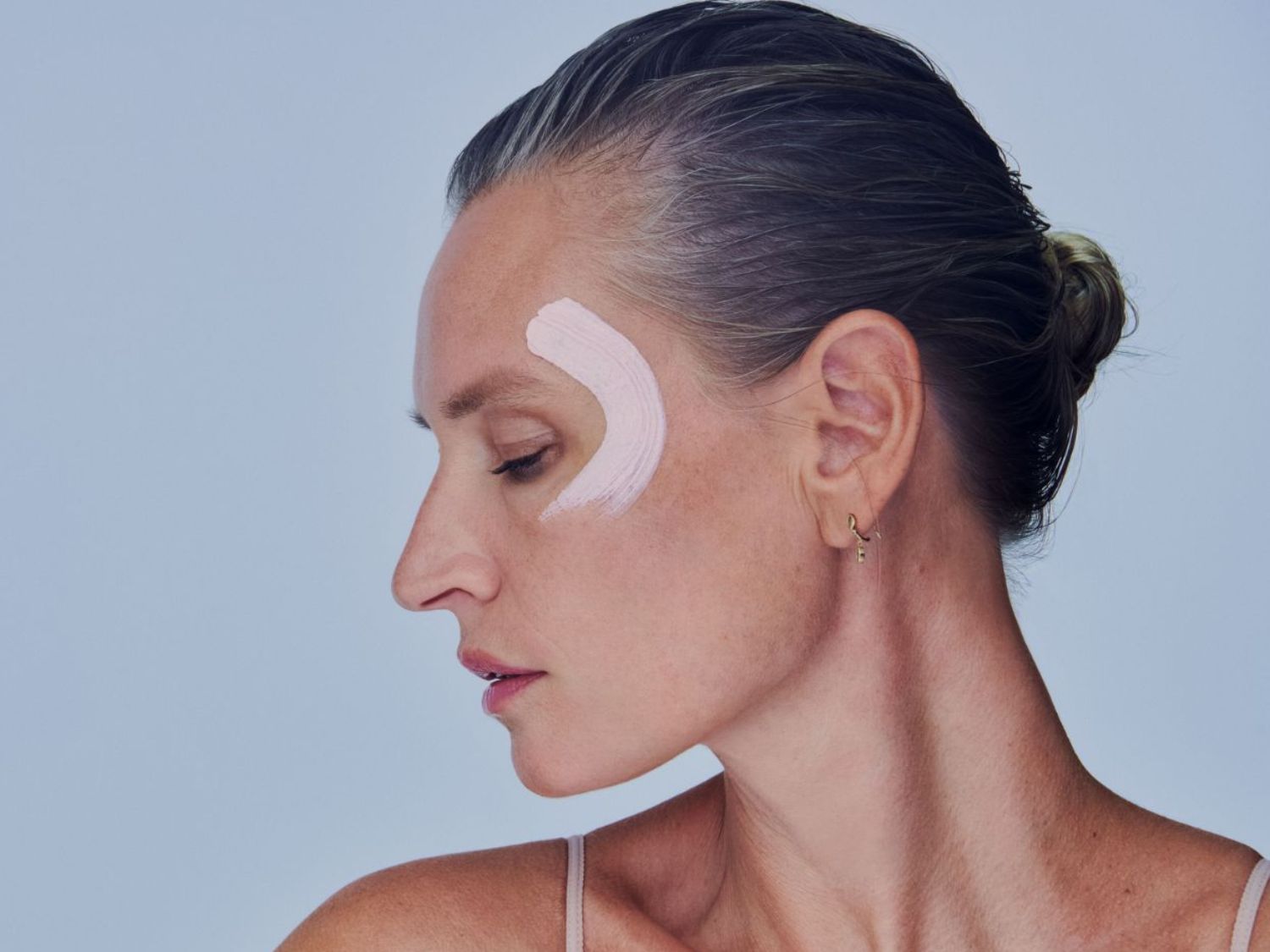 Profil d'une femme avec une touche de crème rose en forme de croissant sur le visage, peau claire et cheveux attachés.
