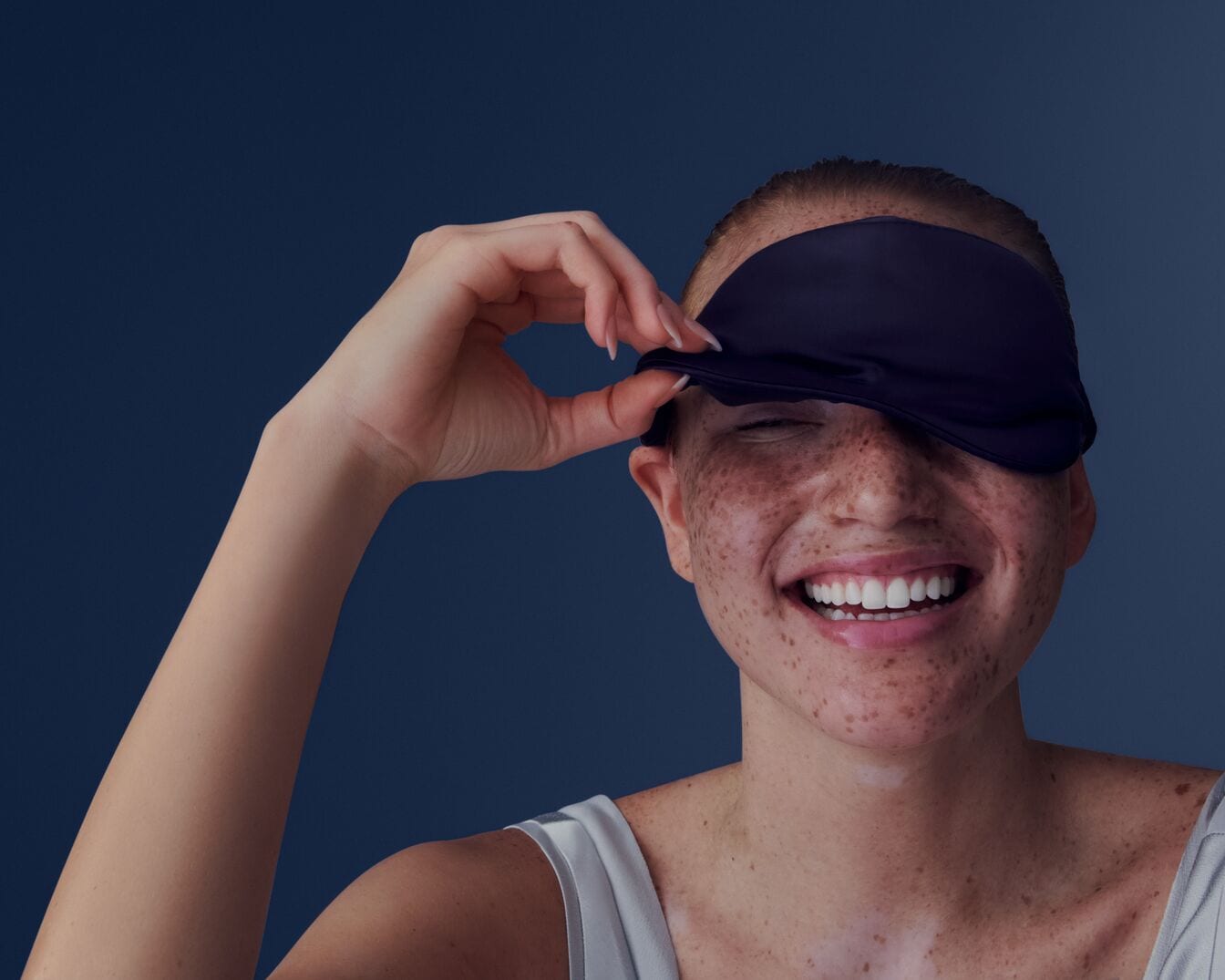 Eine lächelnde Frau mit Sommersprossen hebt eine marineblaue Schlafmaske von ihren Augen.