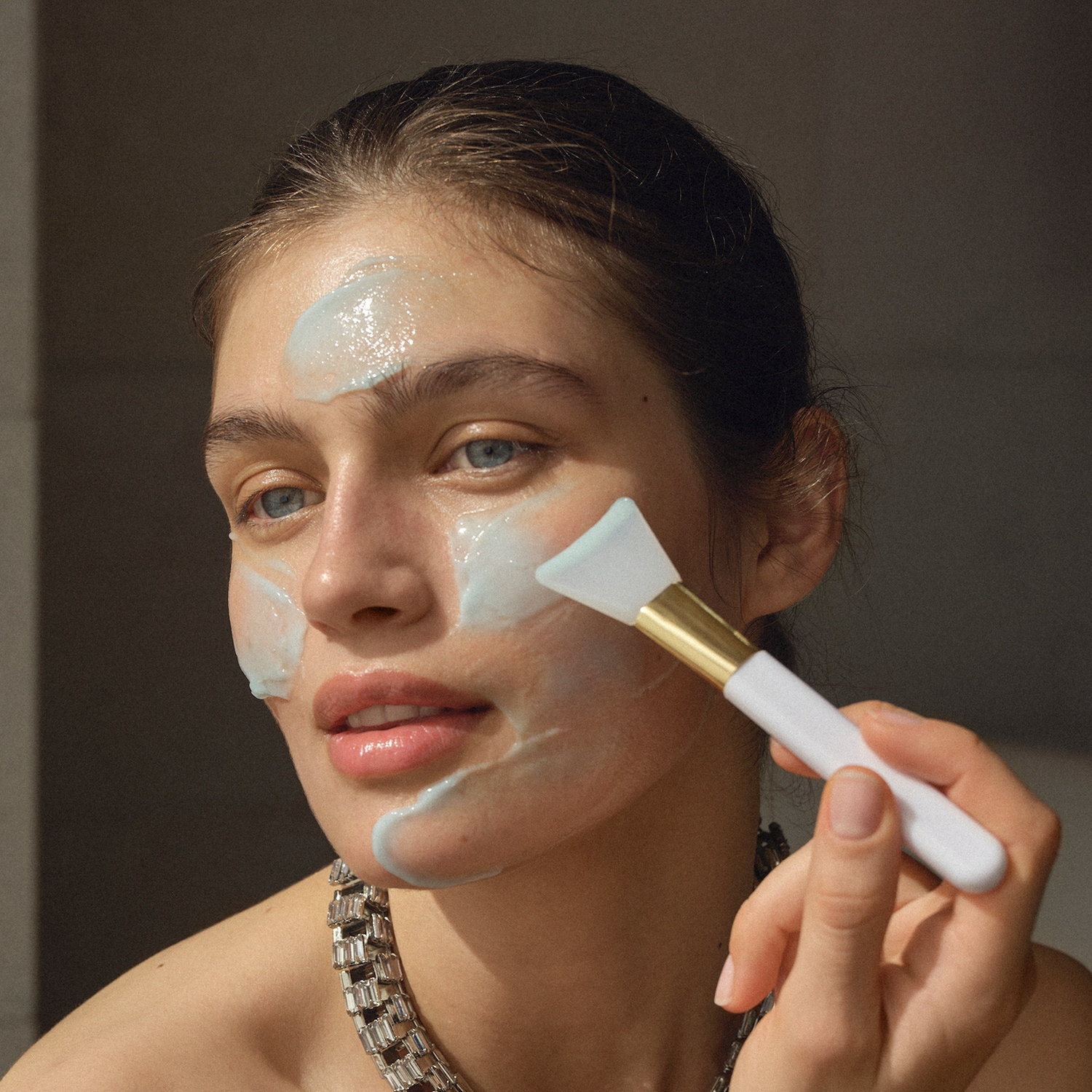 Une femme applique un masque bleu clair sur son visage avec un pinceau blanc et doré, pour une peau éclatante et soignée.