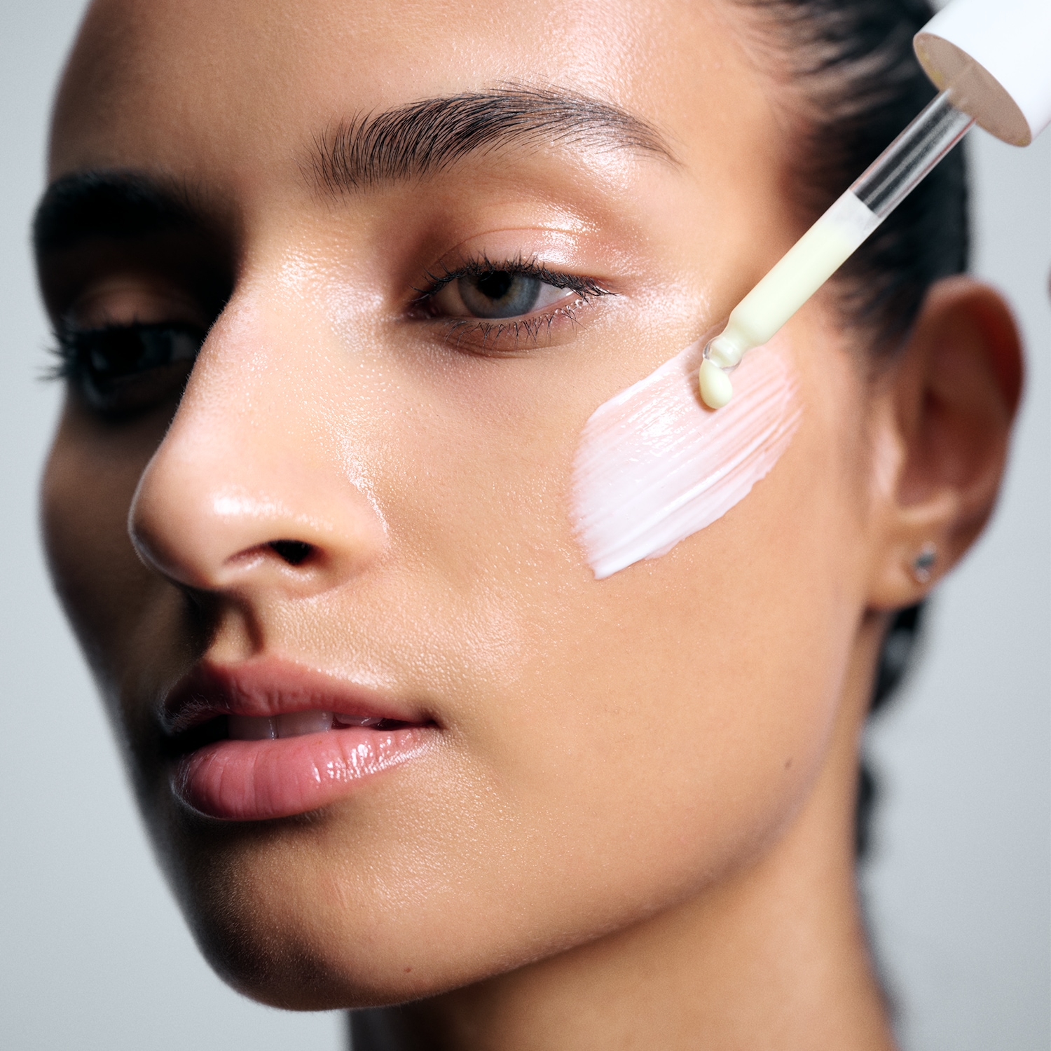 Une femme applique un sérum blanc laiteux sur sa joue avec une pipette, mettant en valeur une peau hydratée et éclatante.