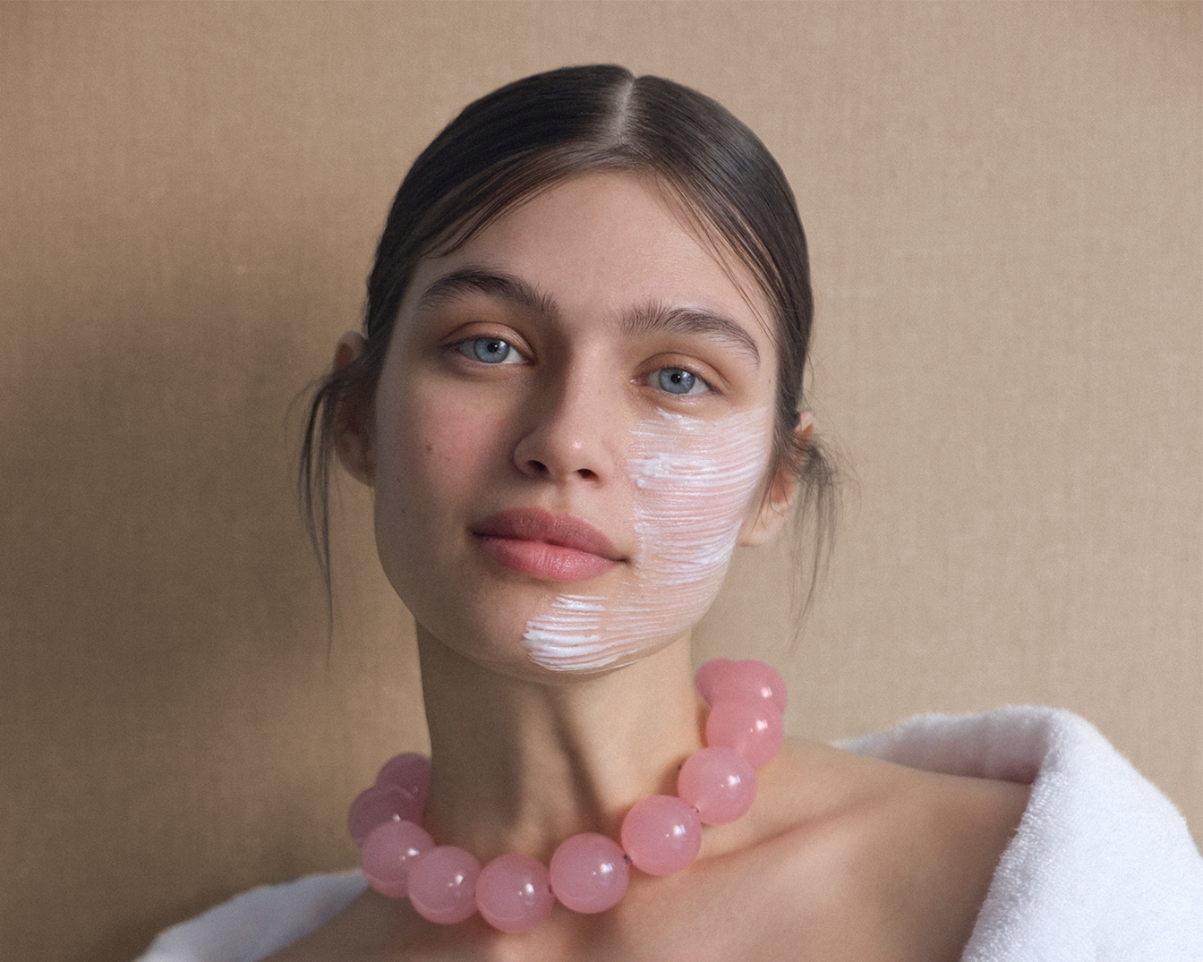 Portrait d'une jeune femme avec un masque blanc sur la moitié du visage, portant un collier de perles roses et une serviette blanche sur les épaules.