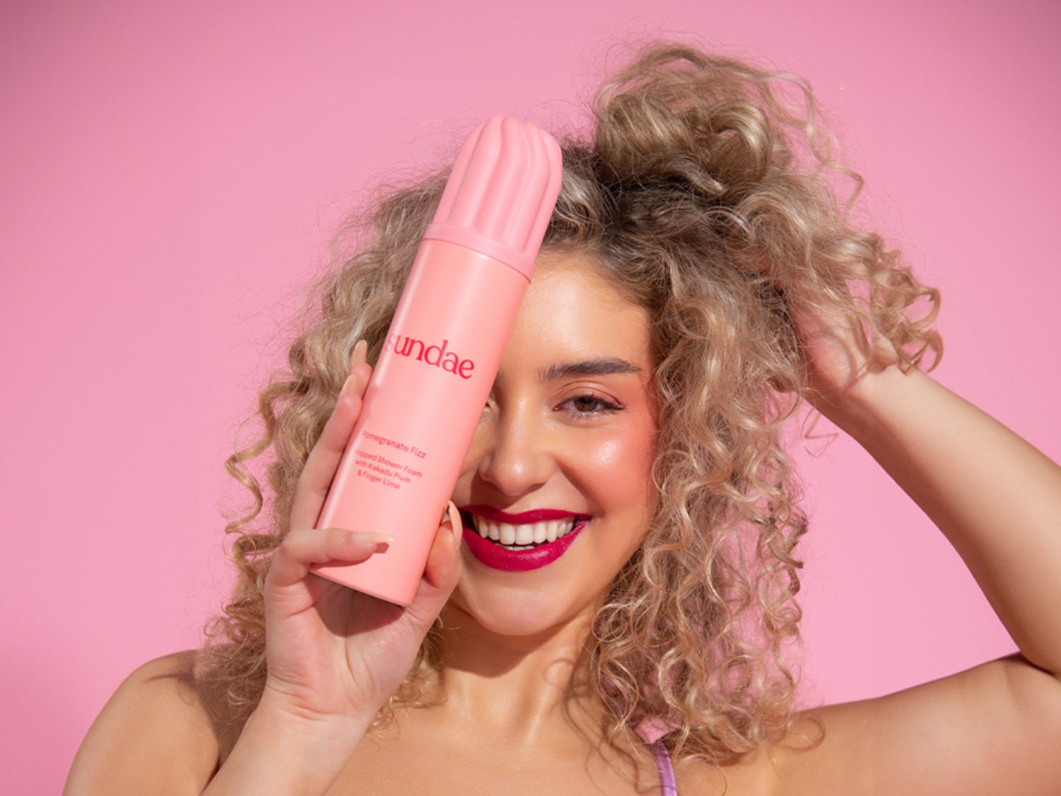 Een lachende vrouw met krullend blond haar houdt een roze Sundae Pomegranate Fizz Whipped Shower Foam bus voor haar gezicht, met de tekst "Pomegranate Fizz Whipped Shower Foam with Kakadu Plum & Finger Lime" duidelijk zichtbaar.