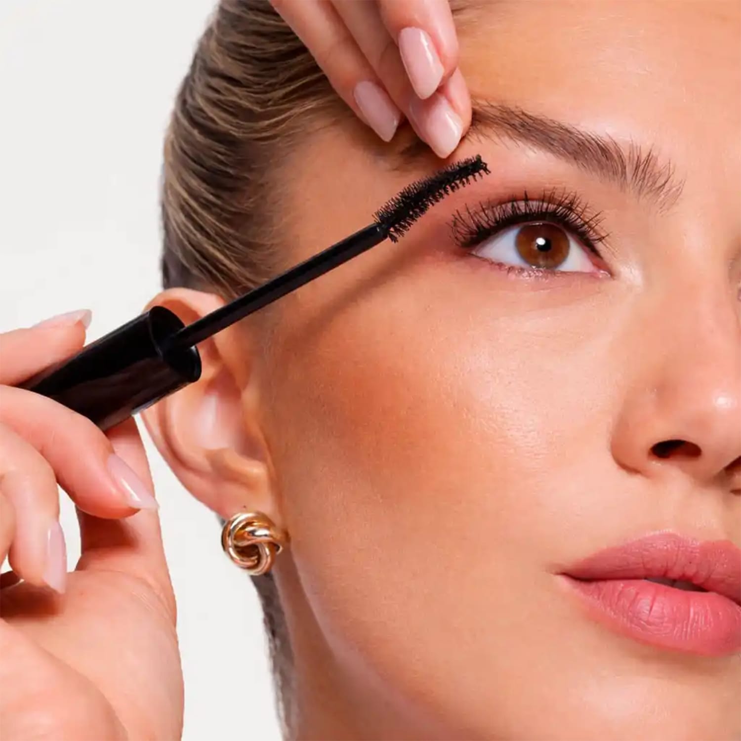 Een vrouw brengt zwarte mascara aan op haar wimpers met een borsteltje, haar gouden oorbel is zichtbaar.