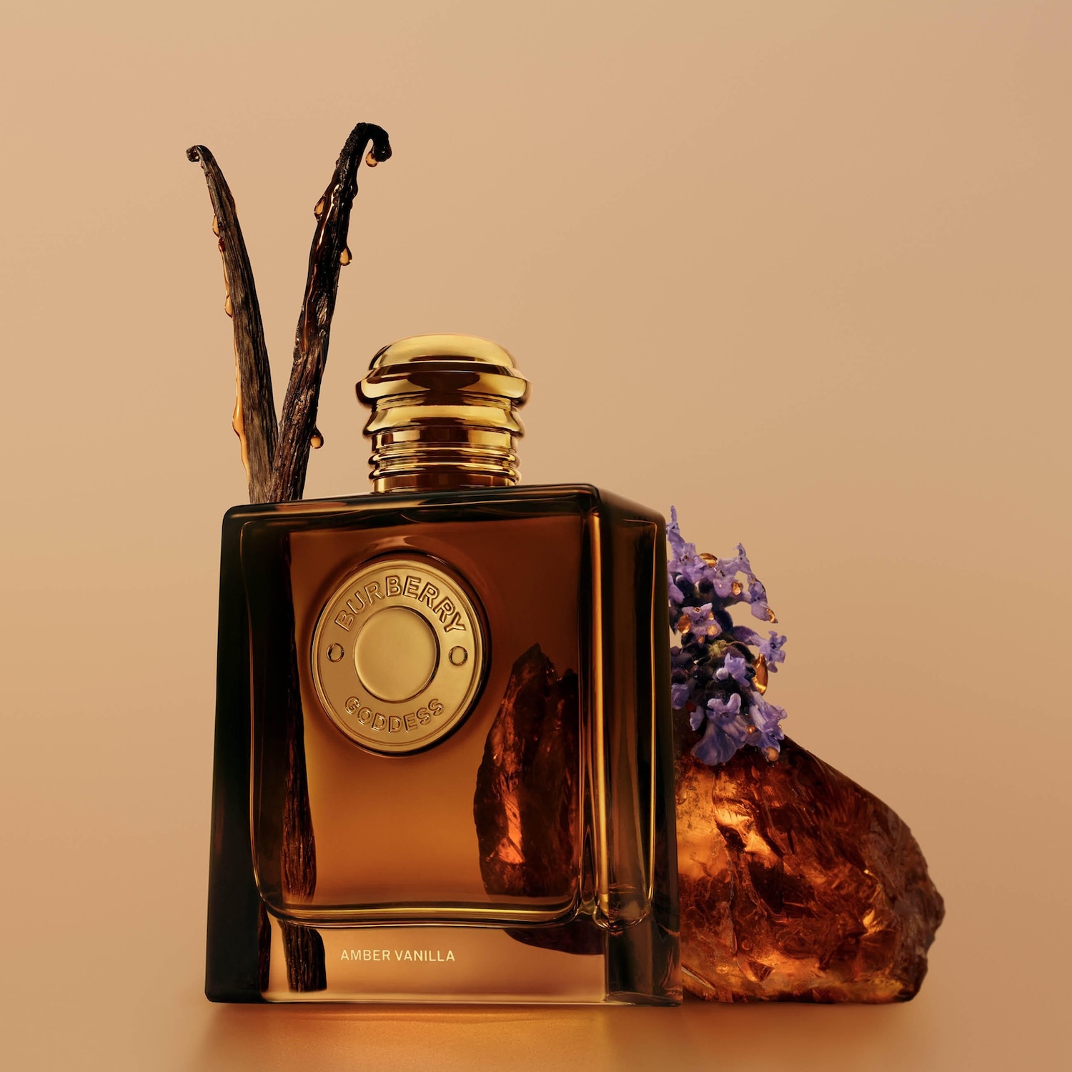 Burberry Goddess Amber Vanilla parfumflesje met gouden dop, omringd door vanillestokjes en een amberkleurige steen met paarse bloemen, op een perzikkleurige achtergrond.
