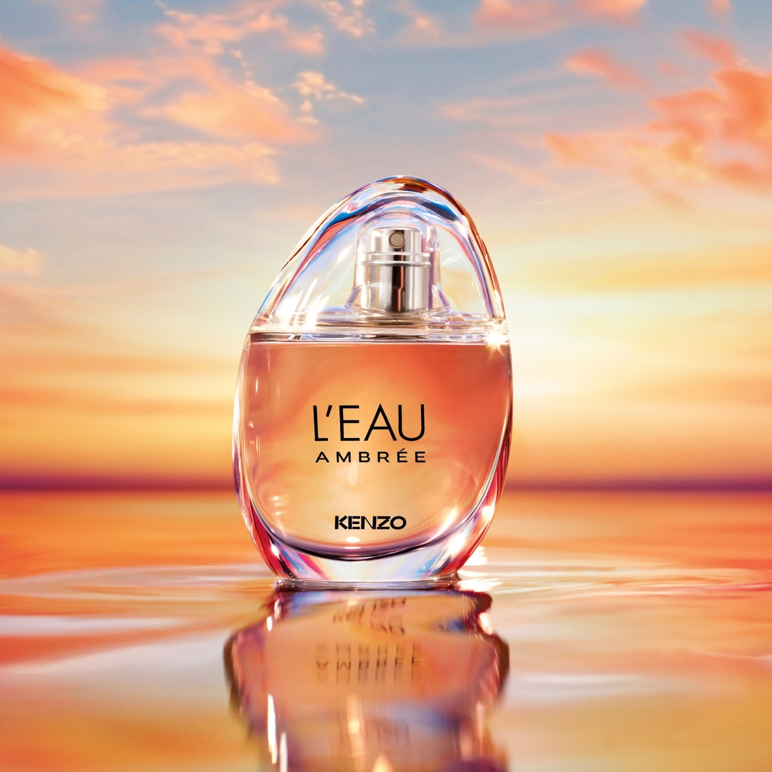 Een heldere glazen fles Kenzo L'Eau Ambrée parfum staat op een reflecterend oppervlak, met een zachte oranje vloeistof binnenin en een achtergrond van een zonsondergang.