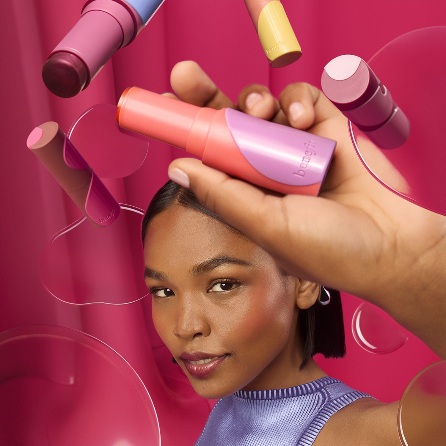 Een vrouw met een paarse top en roze blush houdt een Benefit blush stick vast, omringd door andere blush sticks in roze, perzik en geel.