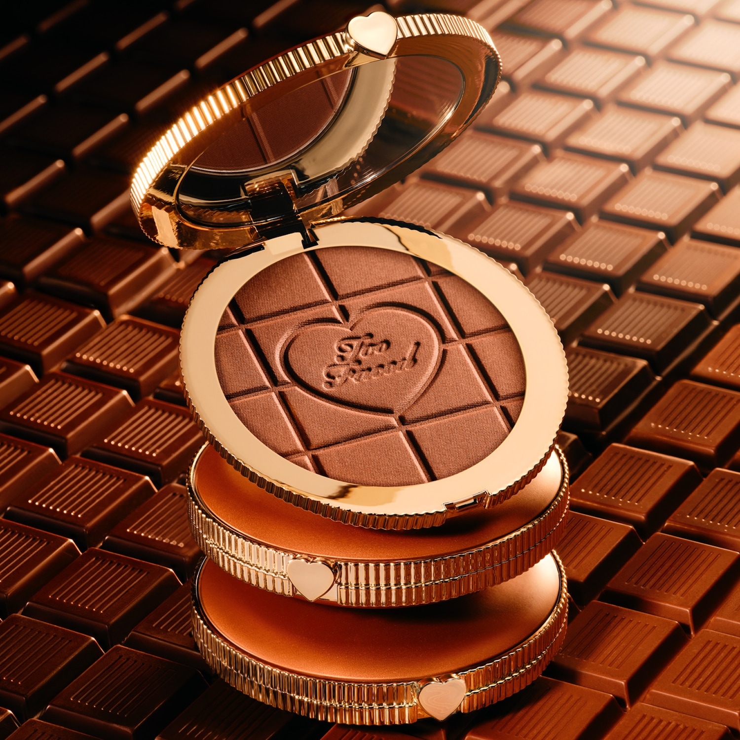 Gouden make-uppoeder van Too Faced, met een chocoladebruine tint en een hartvormig logo, ligt op een achtergrond van chocoladerepen.
