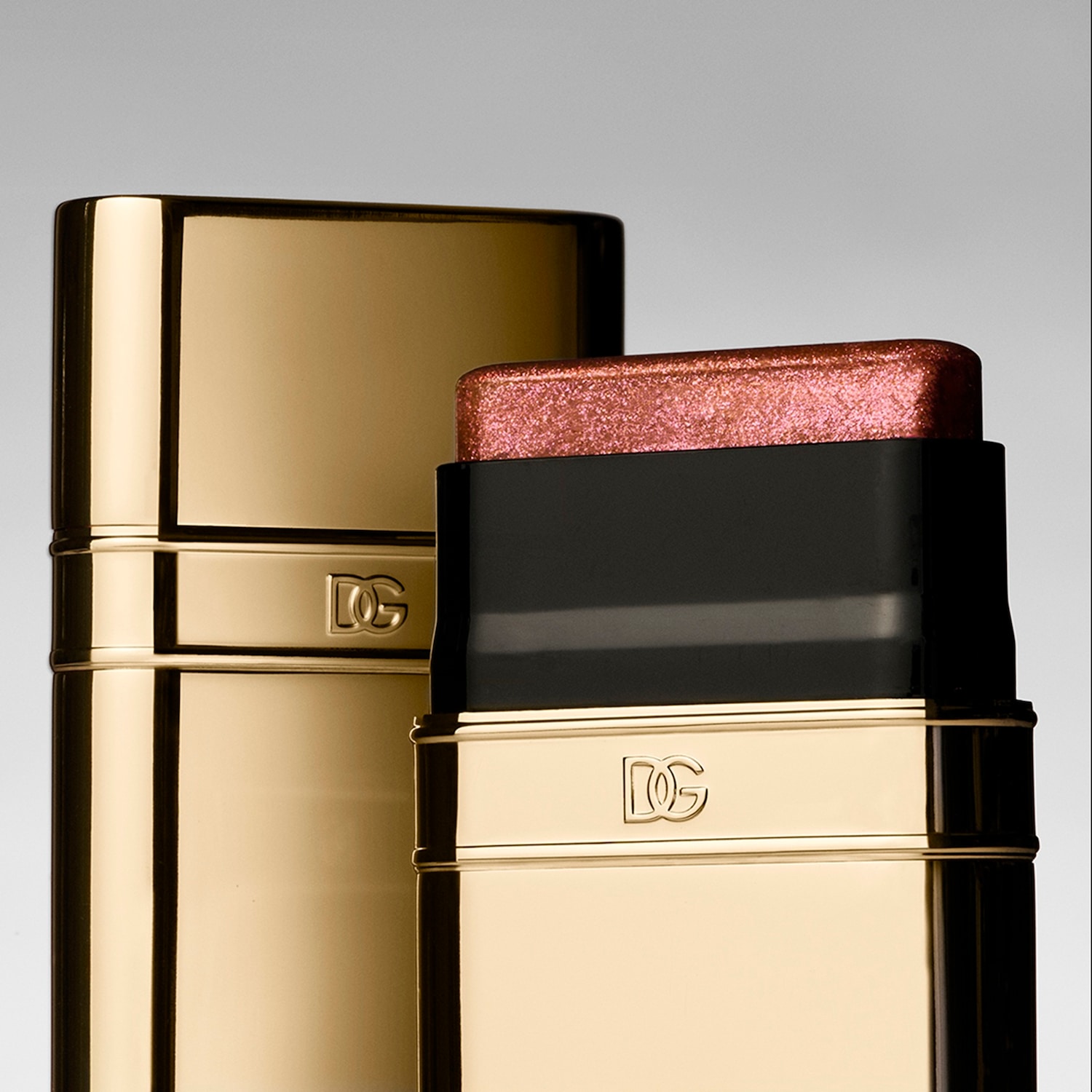 Een close-up van twee Dolce & Gabbana make-up sticks in goudkleurige verpakking, waarvan één geopend is en een glinsterende roze highlighter onthult.