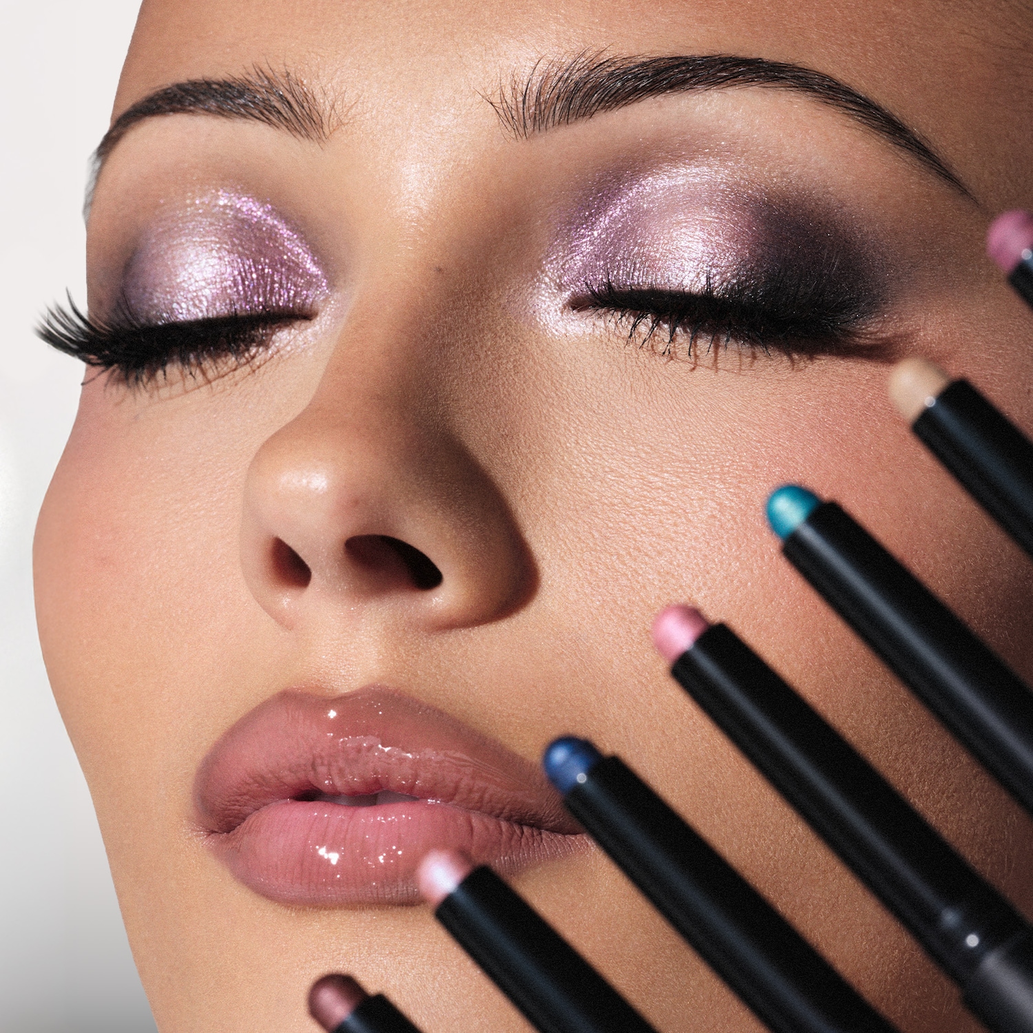 Een close-up van een vrouw met gesloten ogen, met een glanzende lila oogschaduw en glanzende lippen, omringd door oogschaduwsticks in verschillende kleuren.