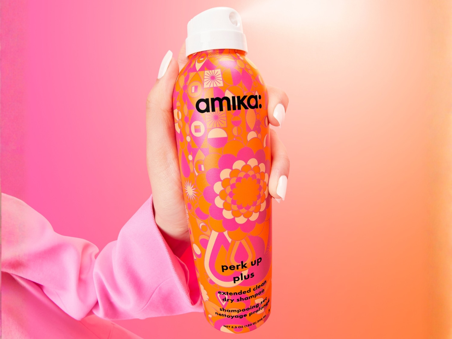 Een hand houdt een kleurrijke fles Amika Perk Up Plus droogshampoo vast, met een bloemmotief in roze en oranje tegen een roze achtergrond.