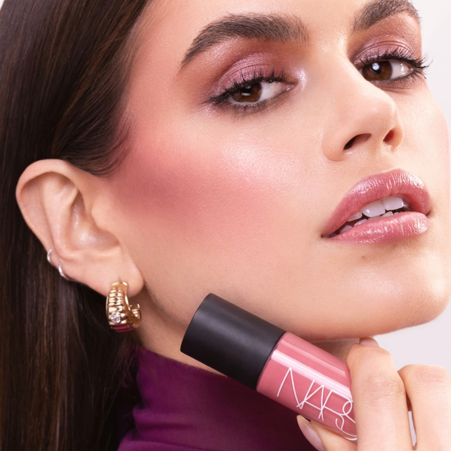 Close-up van een vrouw met roze oogschaduw en blush, die een NARS lipproduct vasthoudt, wat de zachte en stralende make-up look benadrukt.