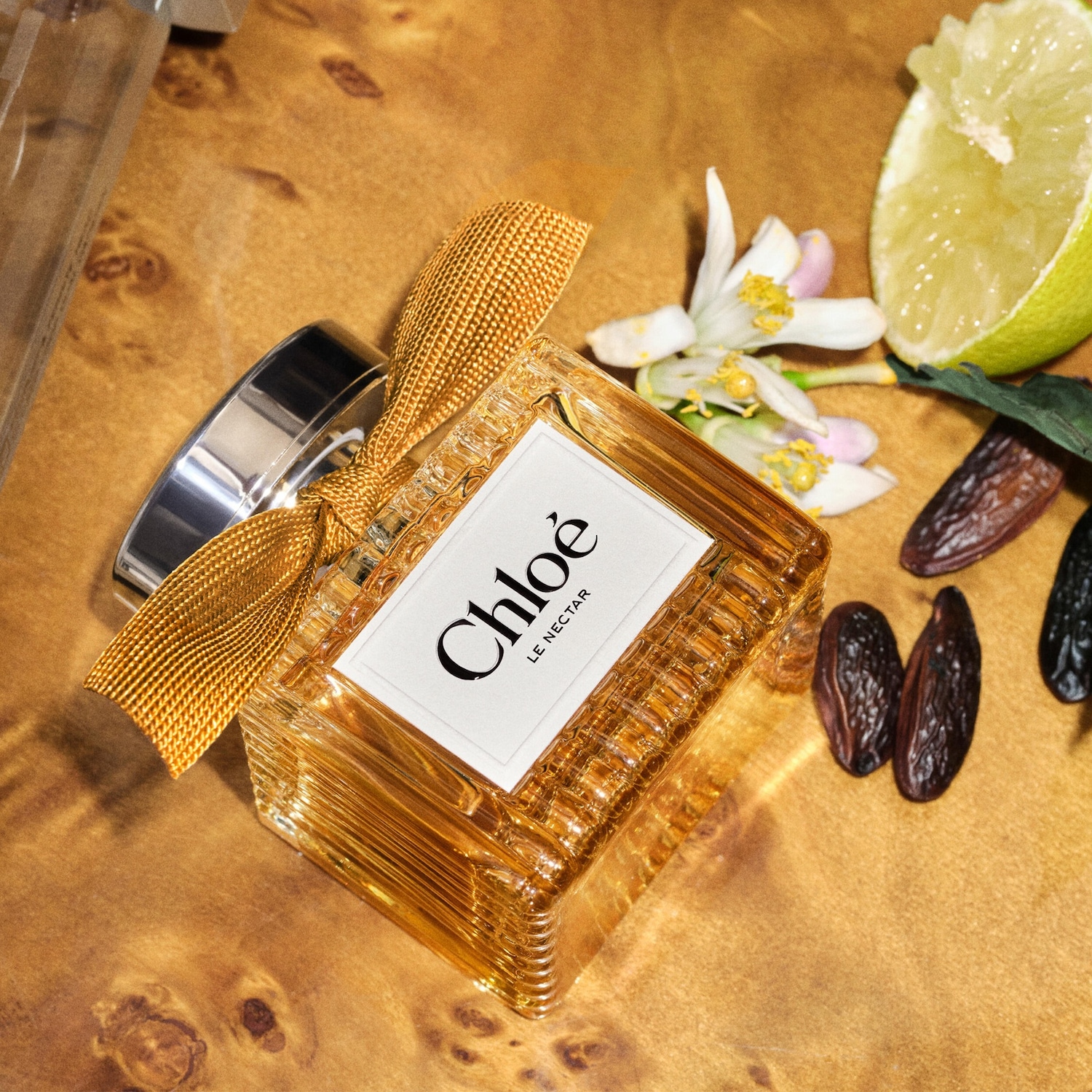Een Chloé Le Nectar parfumflesje met een gouden lint, omringd door witte bloemen, een halve limoen en gedroogde dadels op een houten ondergrond.