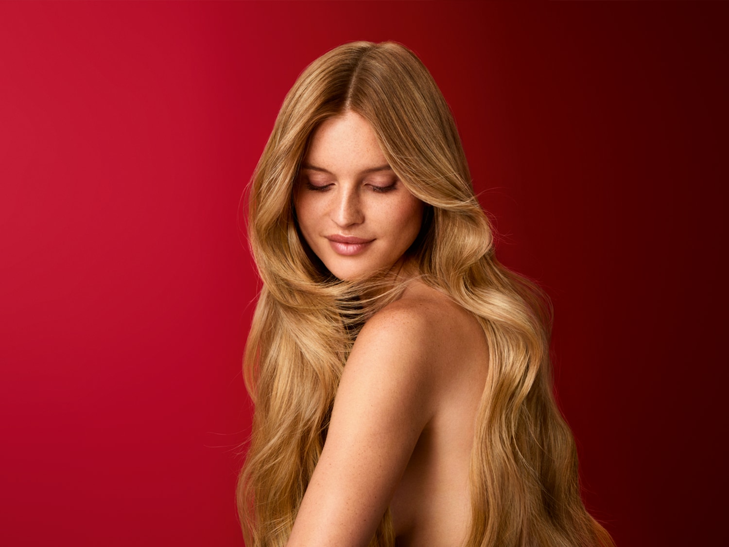 Een vrouw met lang, golvend blond haar en sproeten op haar schouders en gezicht, kijkt naar beneden met gesloten ogen, tegen een rode achtergrond.