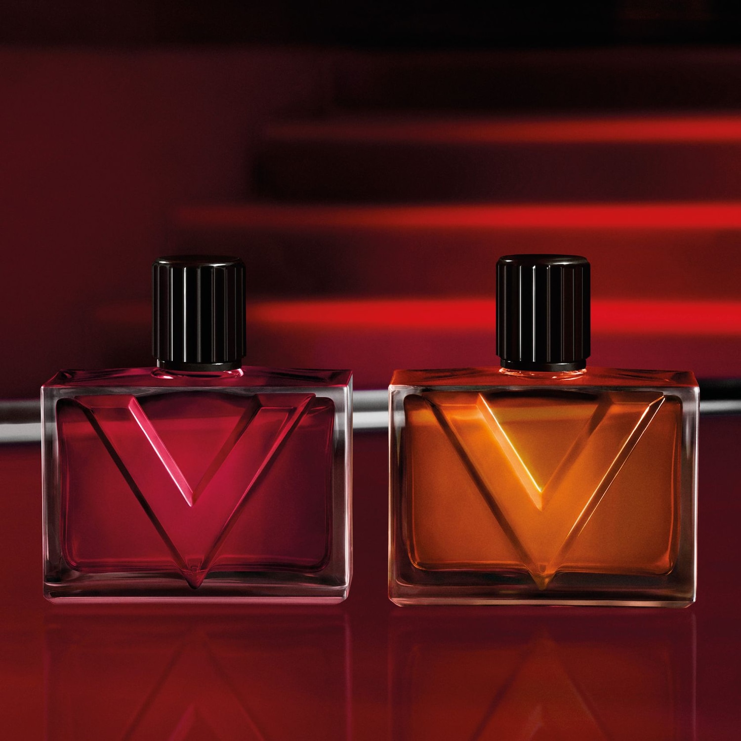 Twee vierkante parfumflesjes met zwarte geribbelde doppen staan naast elkaar, de linker gevuld met een dieprode vloeistof en de rechter met een amberkleurige vloeistof, beide met een verzonken 'V'-vorm aan de voorkant.