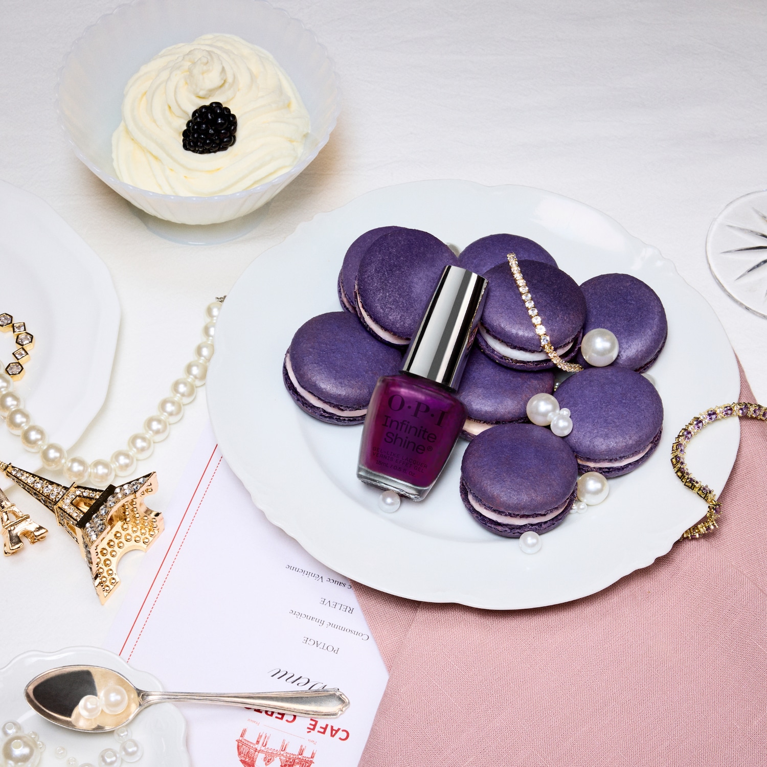 Een OPI Infinite Shine nagellakflesje in een dieppaarse tint staat op een witte schaal met paarse macarons, omringd door parels en sprankelende sieraden, met een toef slagroom en een braam op de achtergrond.