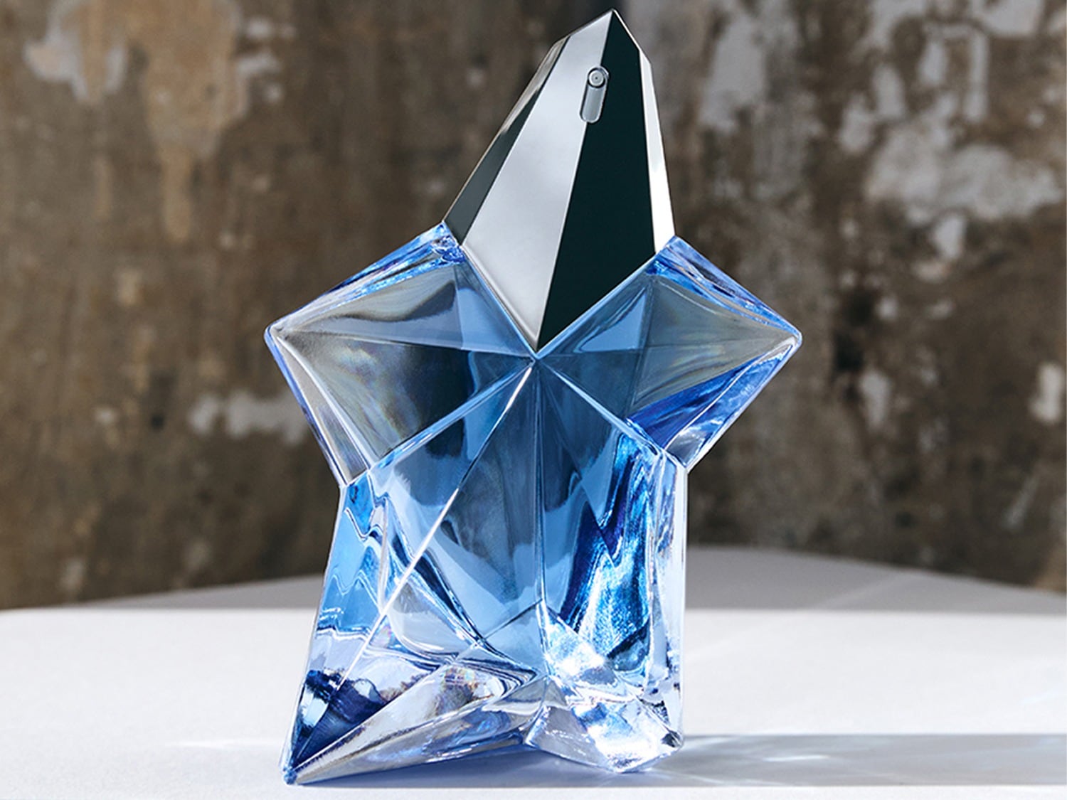 Le parfum Angel de Mugler est présenté dans un flacon en forme d'étoile bleu clair, avec un capuchon argenté brillant, posé sur une surface blanche devant un mur texturé.
