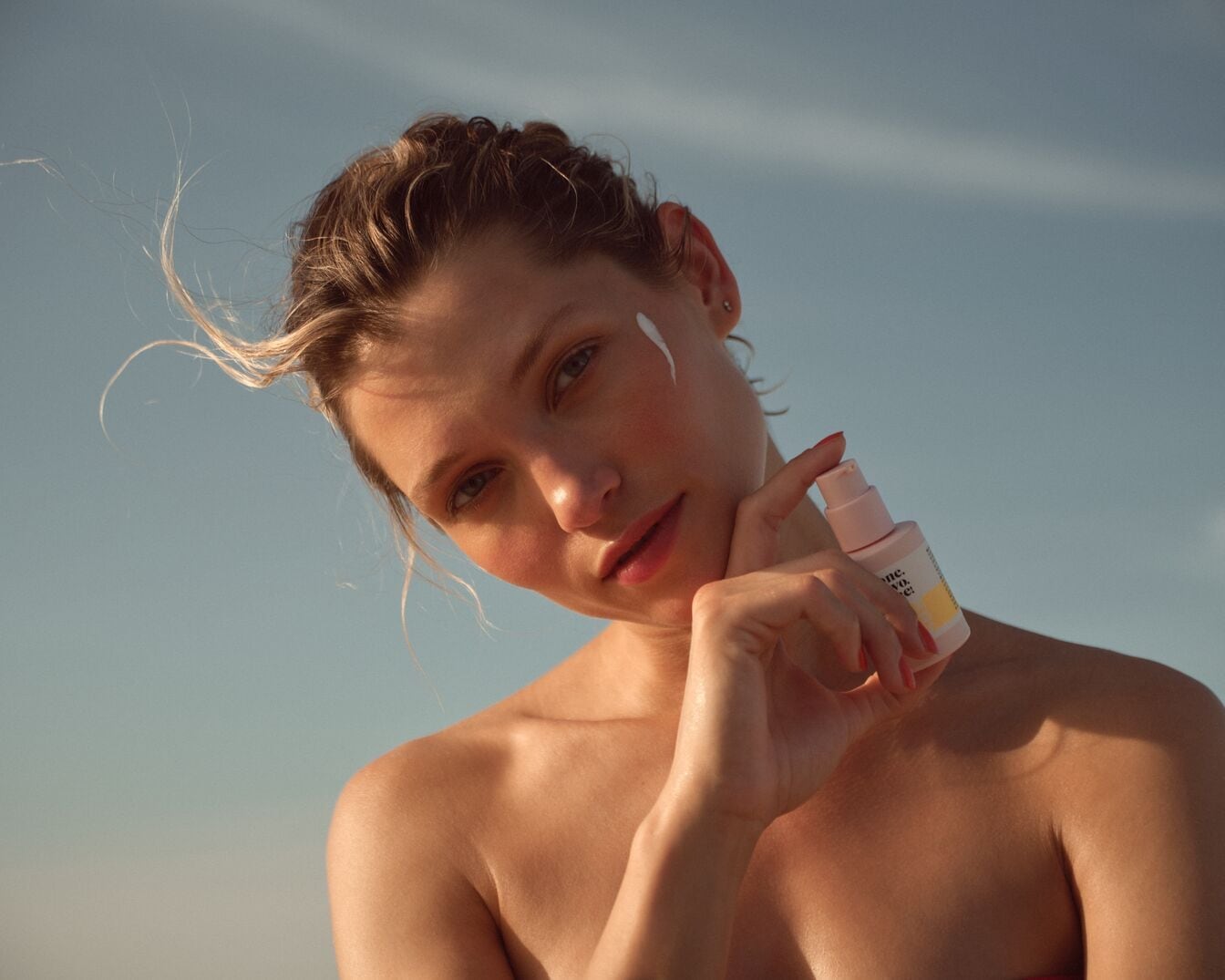 Na slnkom zaliatej pleti modelky sa vyníma ružová fľaštička s krémom, ktorý si nanáša na tvár, čím zdôrazňuje dôležitosť ochrany pred slnkom.