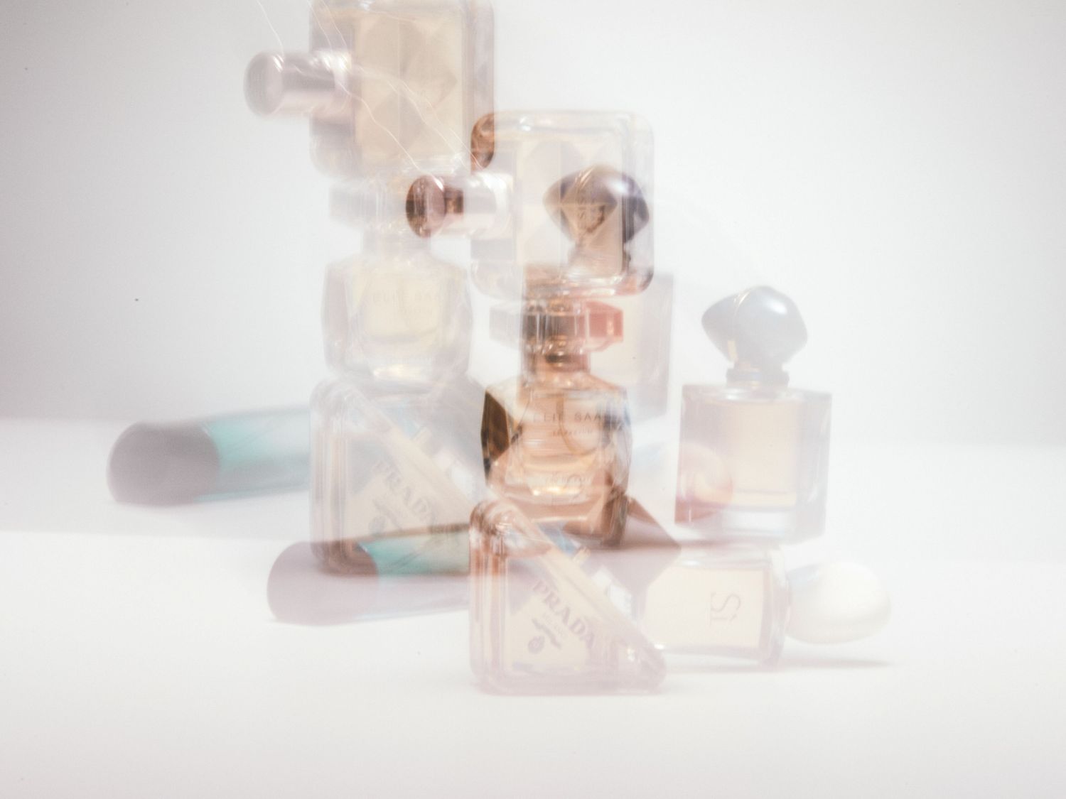 Rozmazané flakony parfémů různých značek, včetně Prada a Elie Saab, na bílém pozadí.