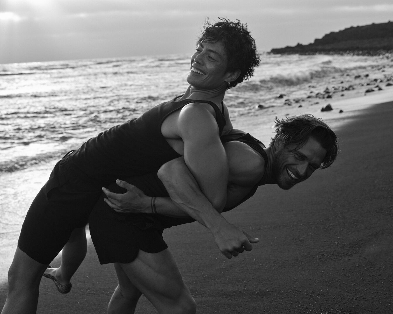 Aufnahme in Schwarzweiß zeigt zwei Männer am Strand, wobei einer den anderen Huckepack trägt und beide mit strahlendem Lächeln die Freude am Zusammensein genießen.