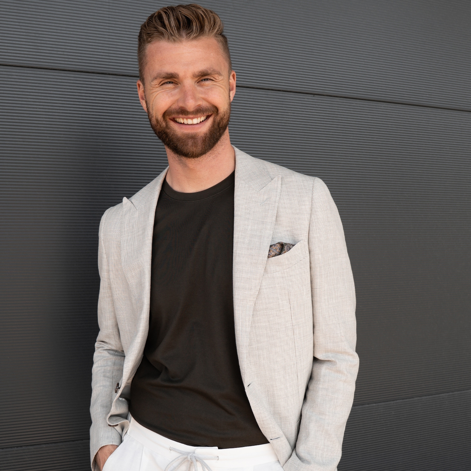 Ein lächelnder Mann mit Bart trägt ein helles Sakko über einem schwarzen T-Shirt und weiße Hosen vor einer grauen Wand.