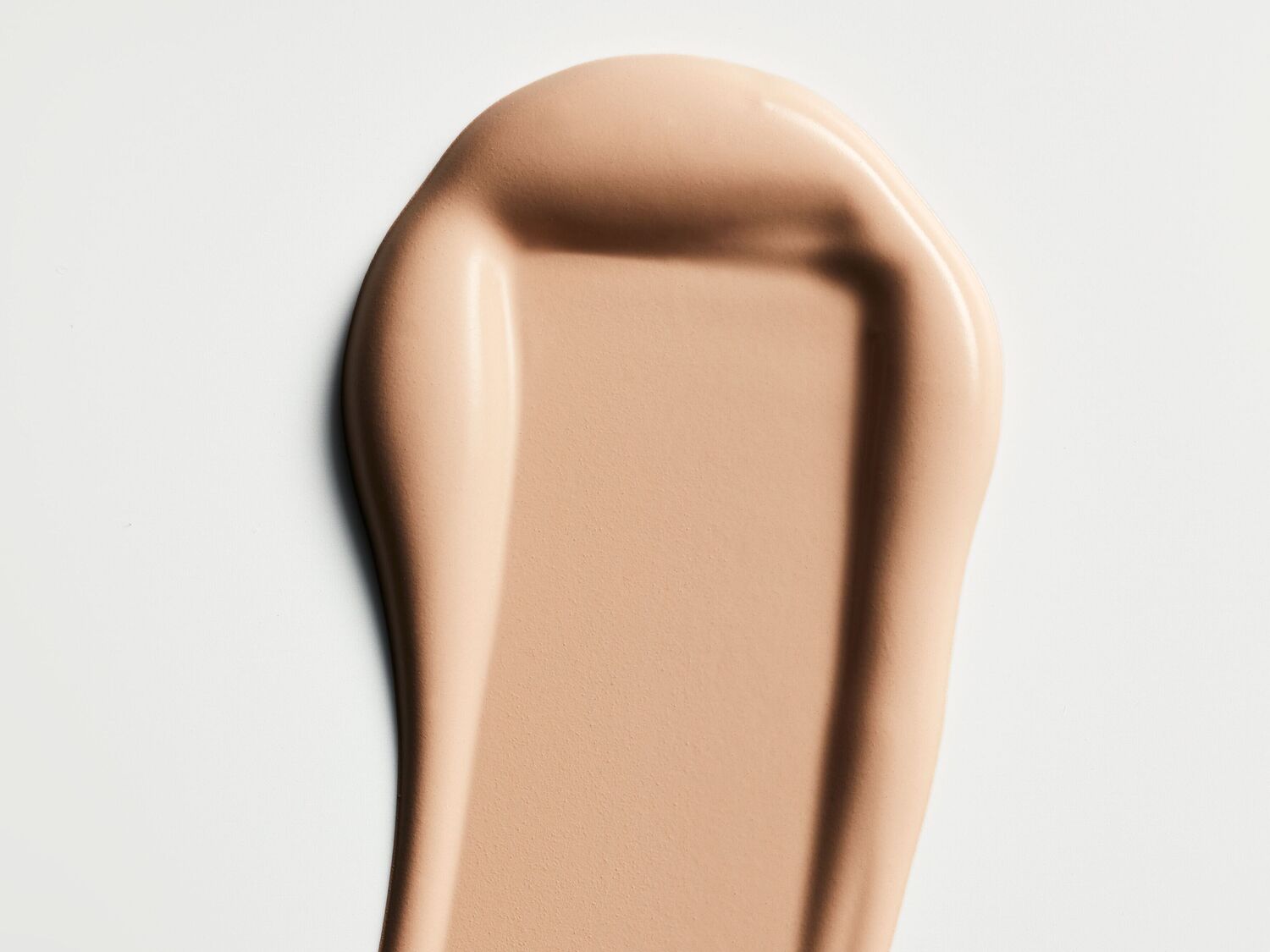 Štai smėlio spalvos kreminės tekstūros kosmetikos priemonės tepinėlis ant balto paviršiaus.