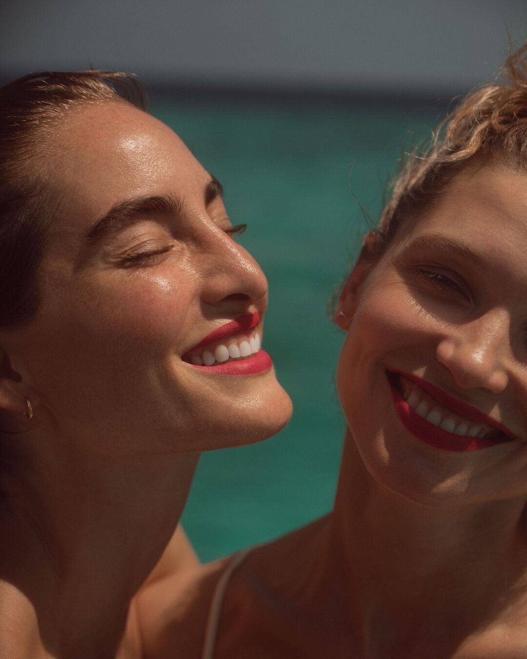 Švytinčios moterys su raudonais lūpų dažais šypsosi priešais akvamarino spalvos foną, spinduliuodamos džiaugsmą ir grožį.