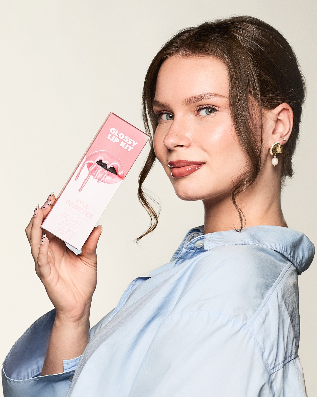 Eine Frau mit hellblauer Bluse hält ein Kylie Cosmetics Glossy Lip Kit in der Hand und präsentiert es.