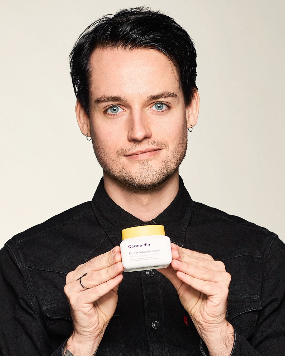 Ein Mann mit blauen Augen und dunklen Haaren hält eine weiße Dose Ceramidin Ectoin-Infused Cream mit gelbem Deckel in den Händen.