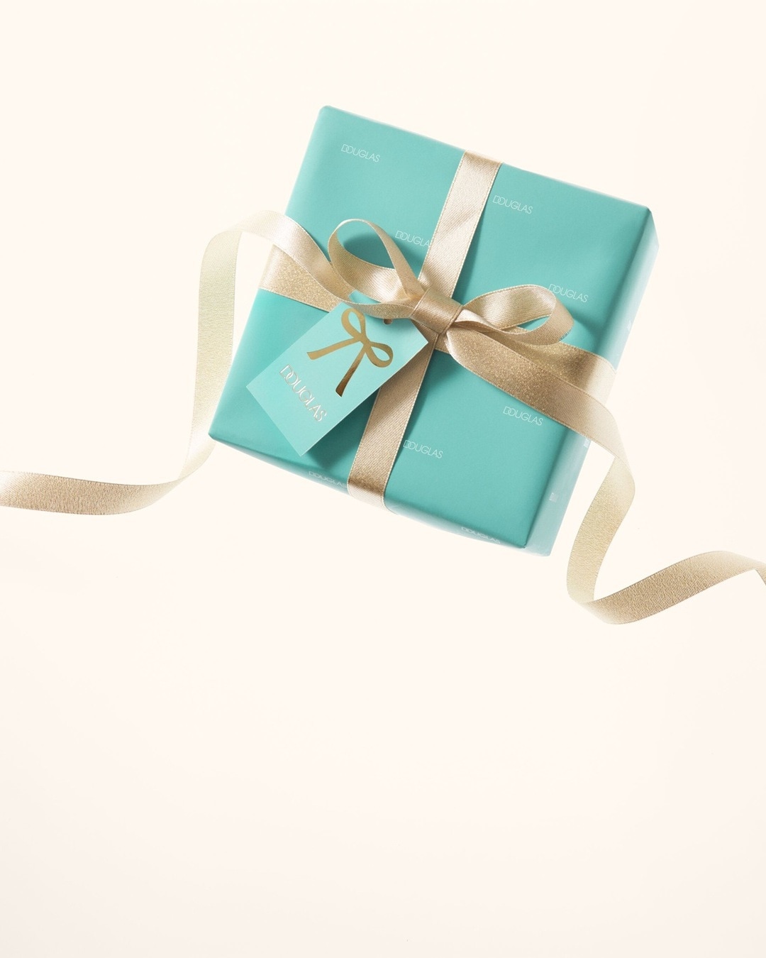 Ein helltürkisfarbenes Geschenk von Douglas, verziert mit einer goldenen Schleife und einem passenden Geschenkanhänger, verspricht Freude.