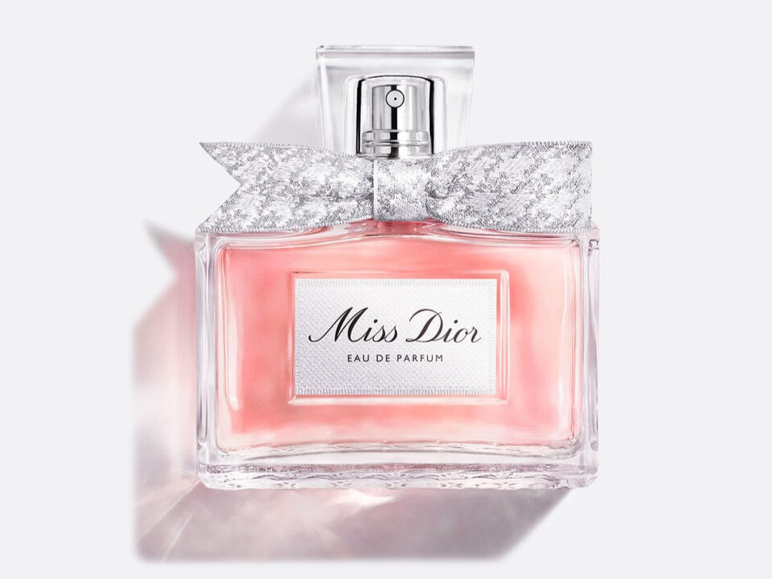 Miss Dior Eau de Parfum in una bottiglia di vetro trasparente con un liquido rosa, adornata con un fiocco argentato e un'etichetta bianca con il nome del profumo in nero.