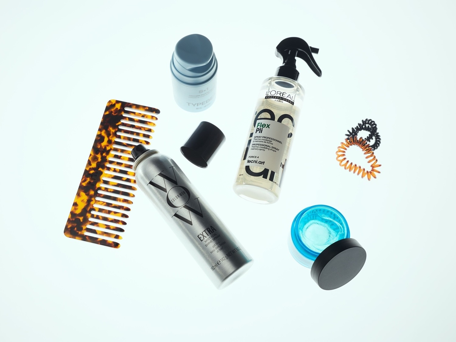 Dargestellt sind verschiedene Haarstyling-Produkte, darunter ein Kamm mit Schildpattmuster, eine silberne Dose COLOR WOW Extra Mist-ical Shine Spray, eine blaue Dose TYPEBE S+1, eine durchsichtige Sprühflasche L'Oréal Professionnel Flex Pli, ein Tiegel mit blauem Inhalt und schwarzem Deckel sowie spiralförmige Haargummis in Schwarz und Orange.