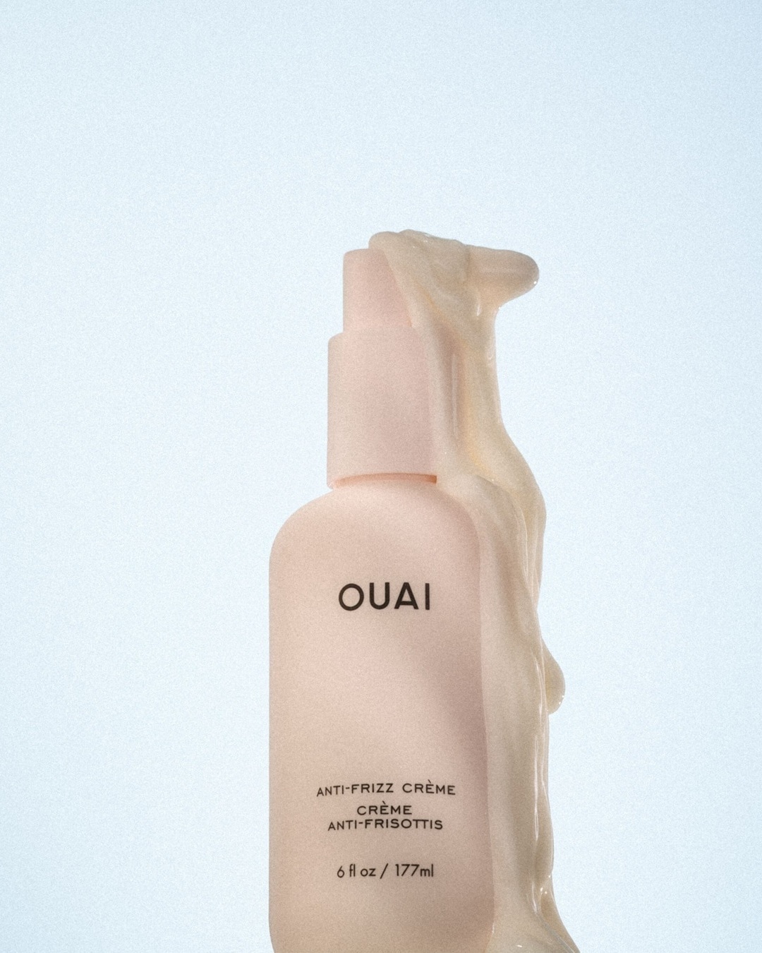 Nahaufnahme der OUAI Anti-Frizz Creme in einer hellrosa Pumpflasche, umgeben von einer cremefarbenen, dicken Textur. Die Flasche verspricht Frizz-Kontrolle.