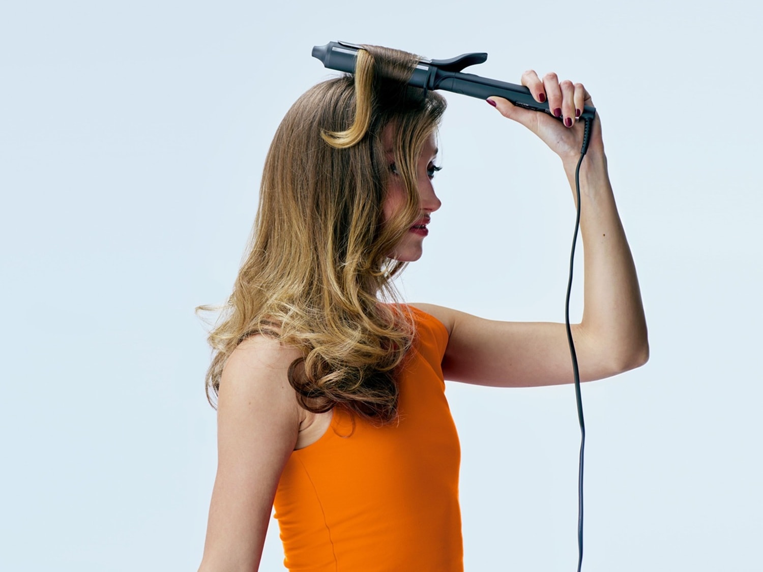 Eine Frau mit orangefarbenem Top lockt ihr blondes Haar mit einem schwarzen Lockenstab.