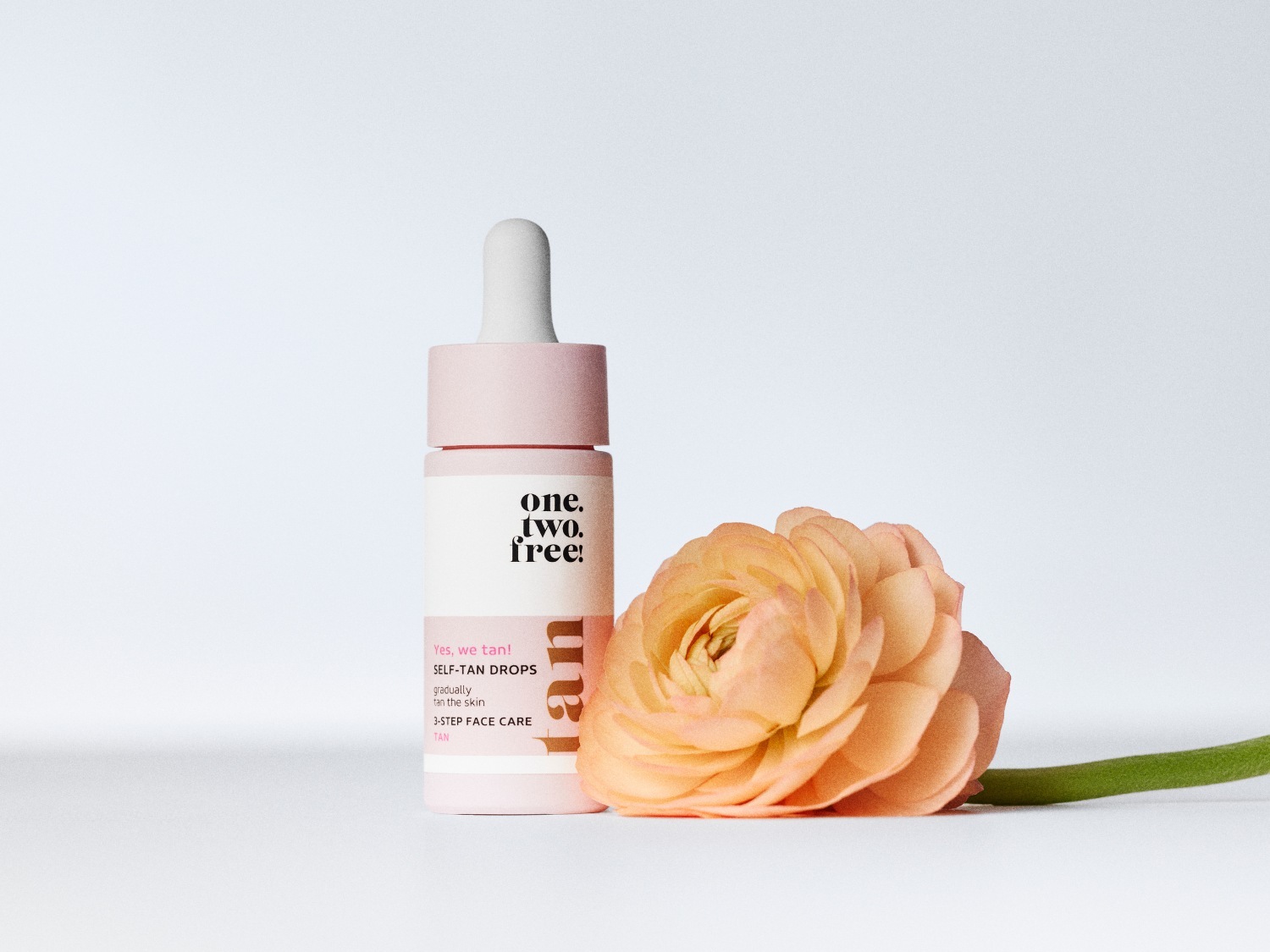 De zelfbruinende druppels van One. Two. Free! in een lichtroze verpakking met een witte pipet, naast een perzikkleurige bloem, beloven een geleidelijke teint voor de huid.