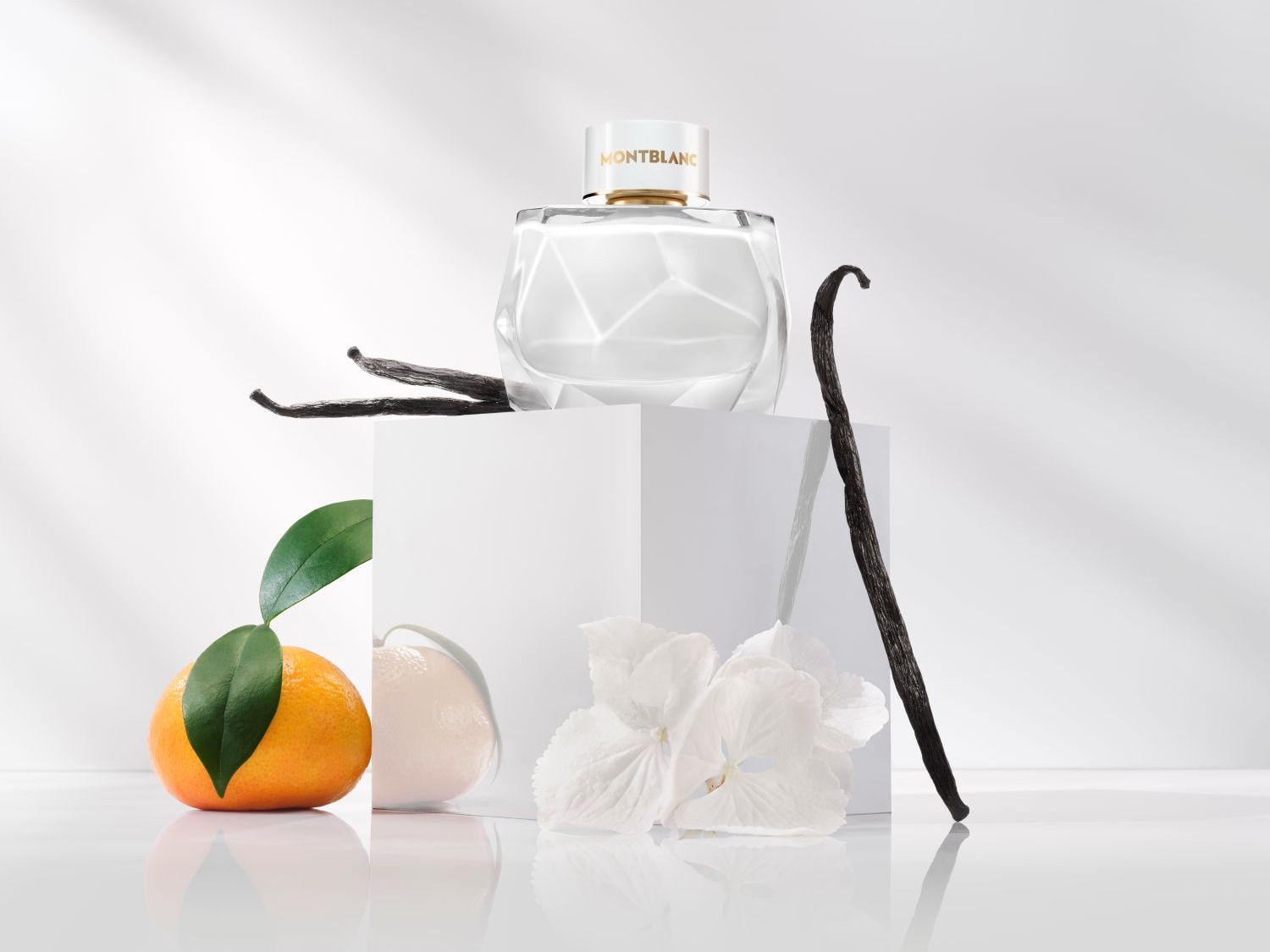 Ein Montblanc-Duft präsentiert sich in einem klaren, facettierten Flakon mit goldenen Akzenten, umgeben von Vanilleschoten, einer Mandarine mit Blättern und einer weißen Blüte.