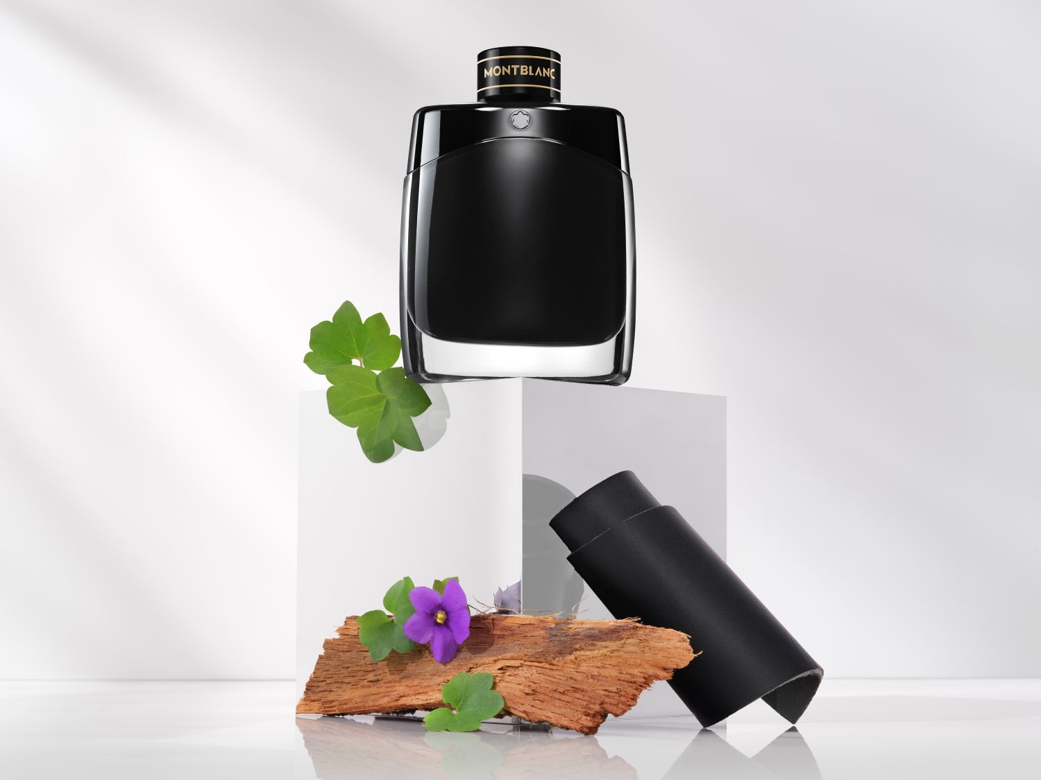 Ein elegantes, schwarzes Montblanc Legend Parfüm steht auf einem weißen Sockel, akzentuiert durch ein Stück Holz, eine violette Blüte, grüne Blätter und ein gerolltes Stück schwarzes Leder.