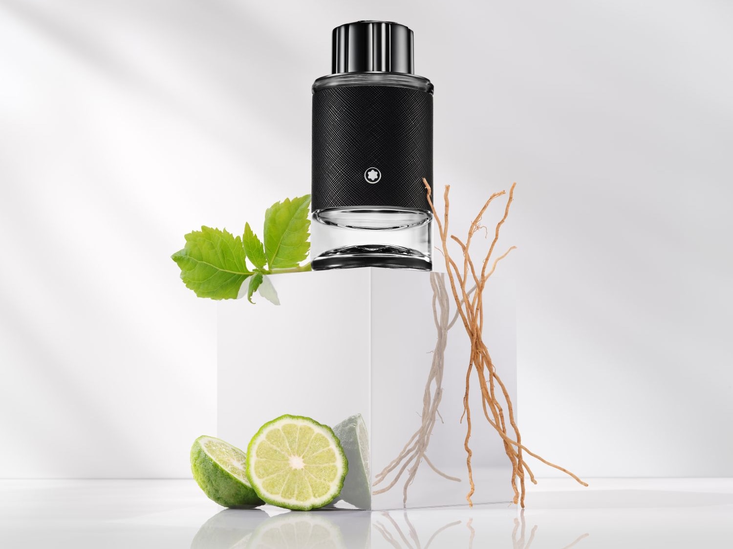Ein Flakon Montblanc-Parfüm mit schwarzem Lederbezug steht auf einem weißen Sockel, umgeben von grünen Blättern, Limettenscheiben und Wurzeln.