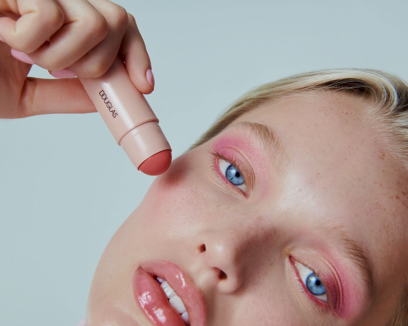 Een vrouw met blauwe ogen en roze oogschaduw houdt een Douglas blush stick vast, klaar om haar wangen een roze gloed te geven.