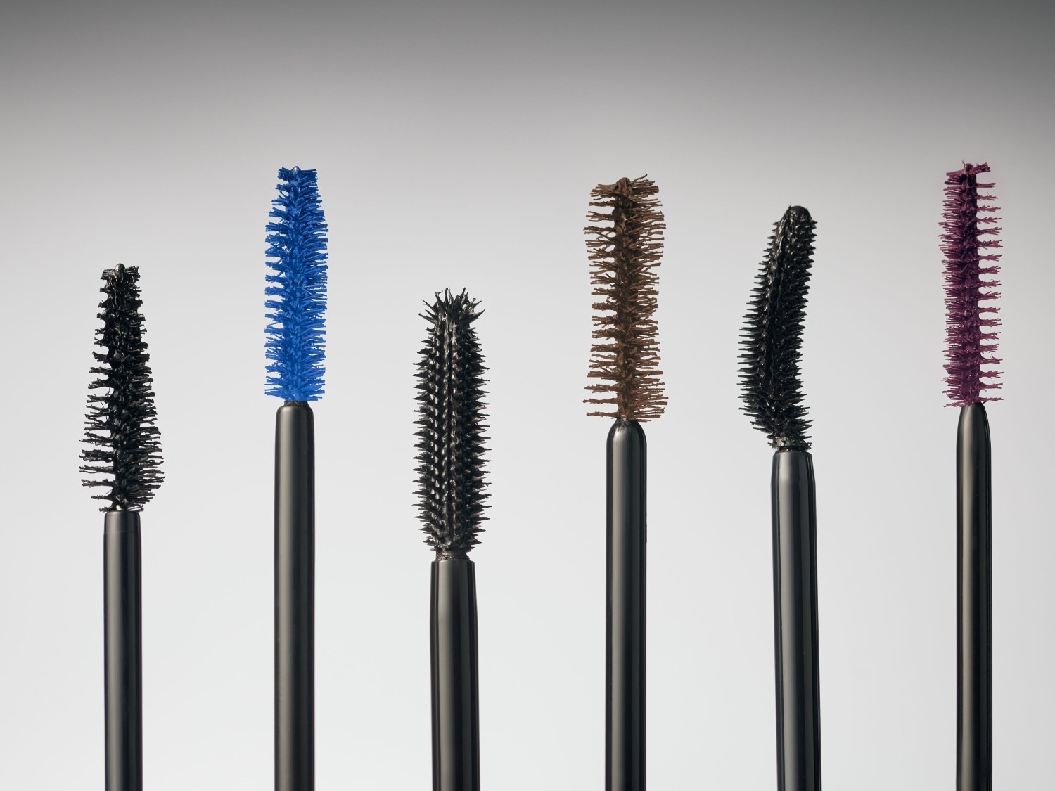 Eine Reihe von Mascara-Bürsten in verschiedenen Farben, darunter Schwarz, Blau, Braun und Lila, die jeweils unterschiedliche Formen und Größen haben.